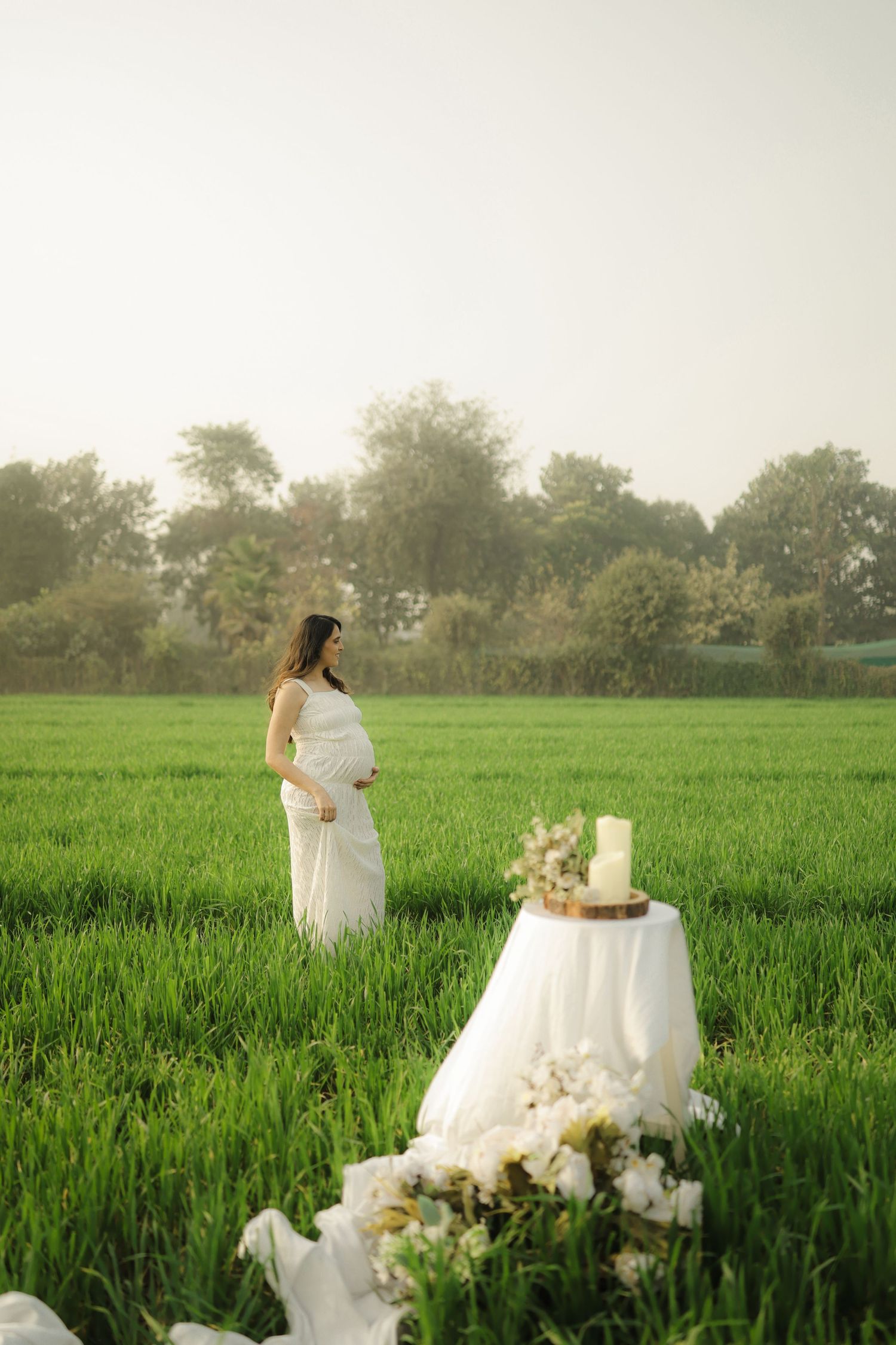 A woman in a flowing white dress poses in a lush green field with a decorated white cake table at sunset.