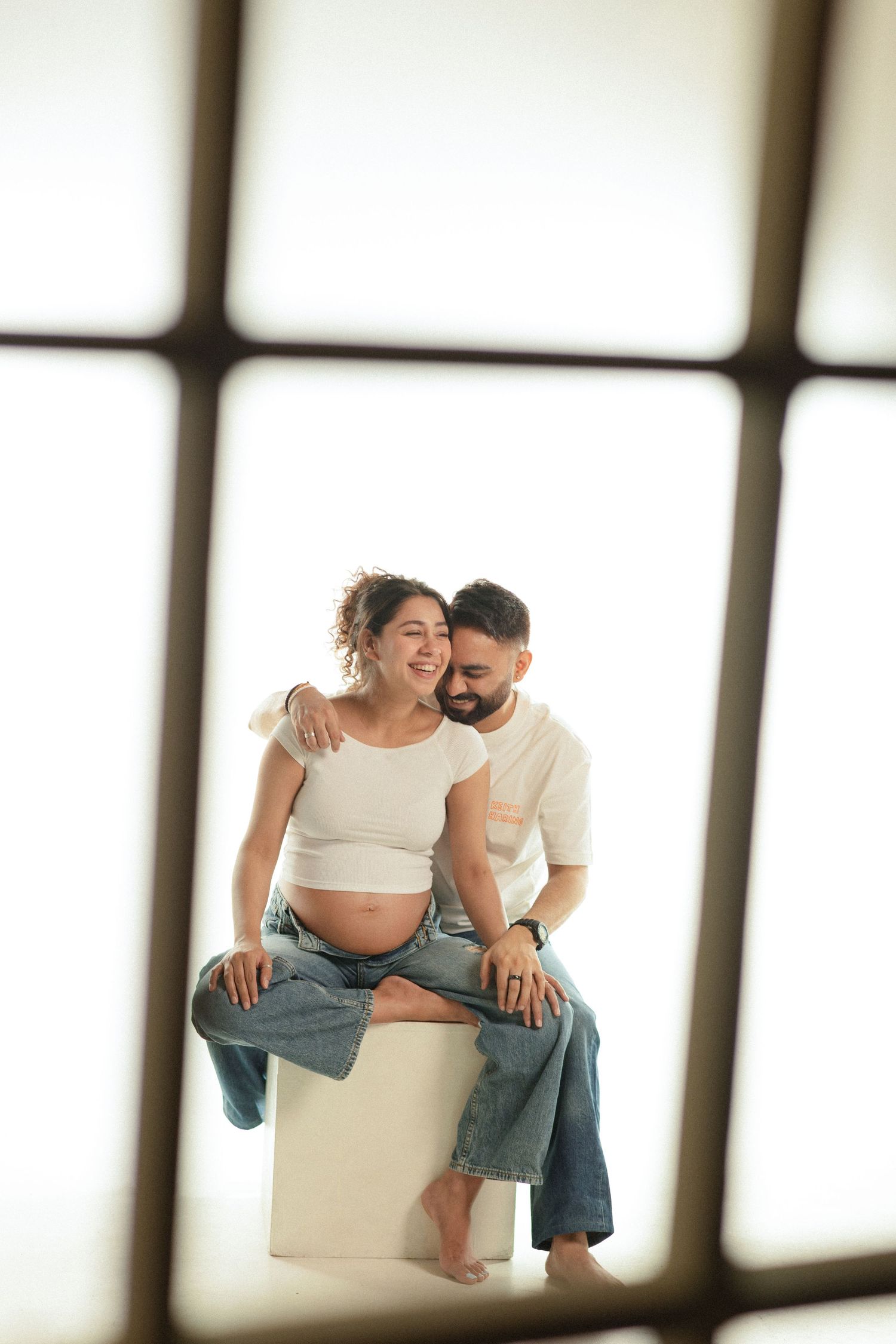 Couple in casual white tops and jeans share intimate moments while sitting on white furniture in sunlit room.