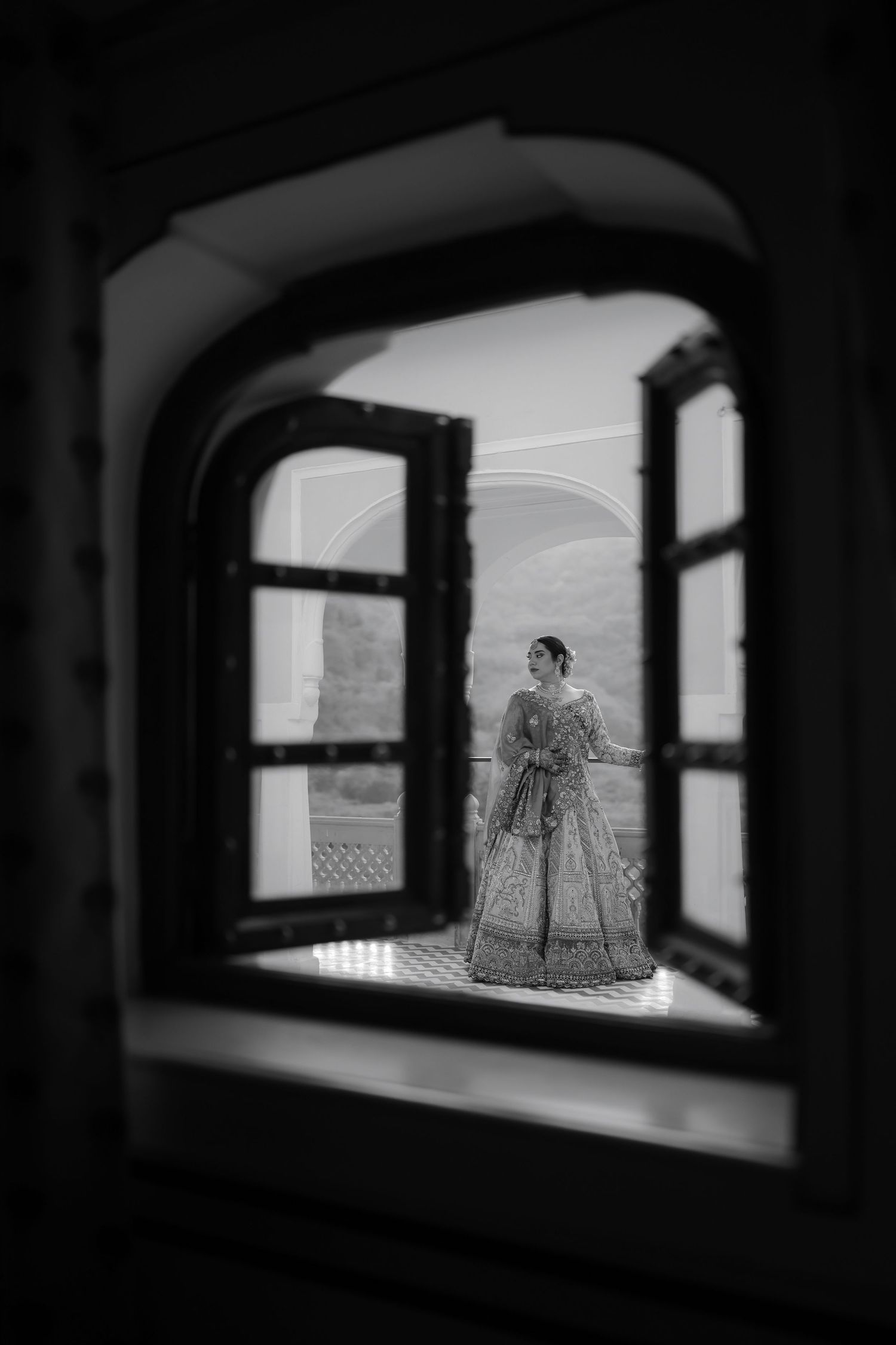 Black and white photo of a bride in lehenga framed by an arched window at a royal Indian palace.
