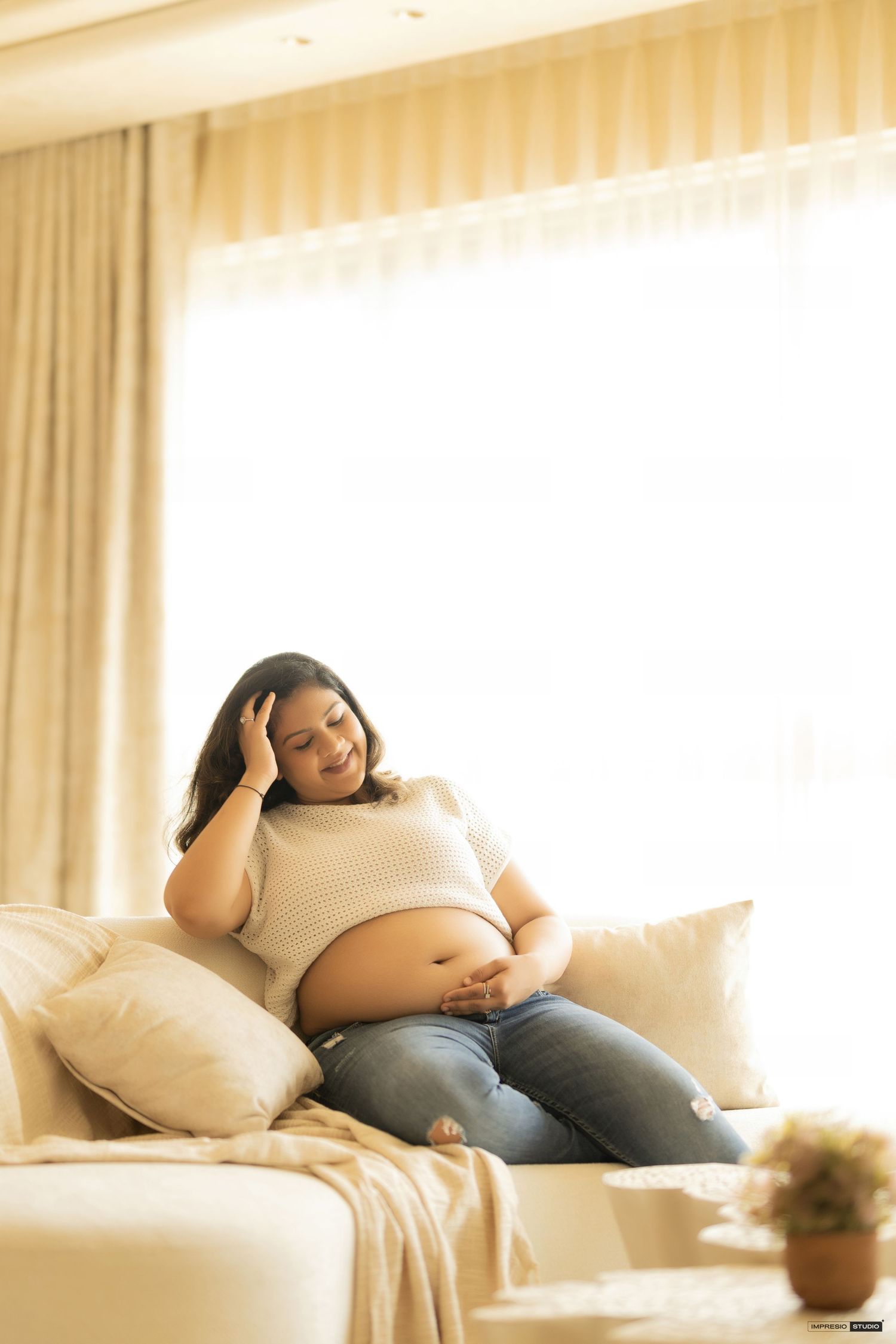 Pregnant woman sitting on sofa touching her belly and head, wearing white top and jeans in bright living room.