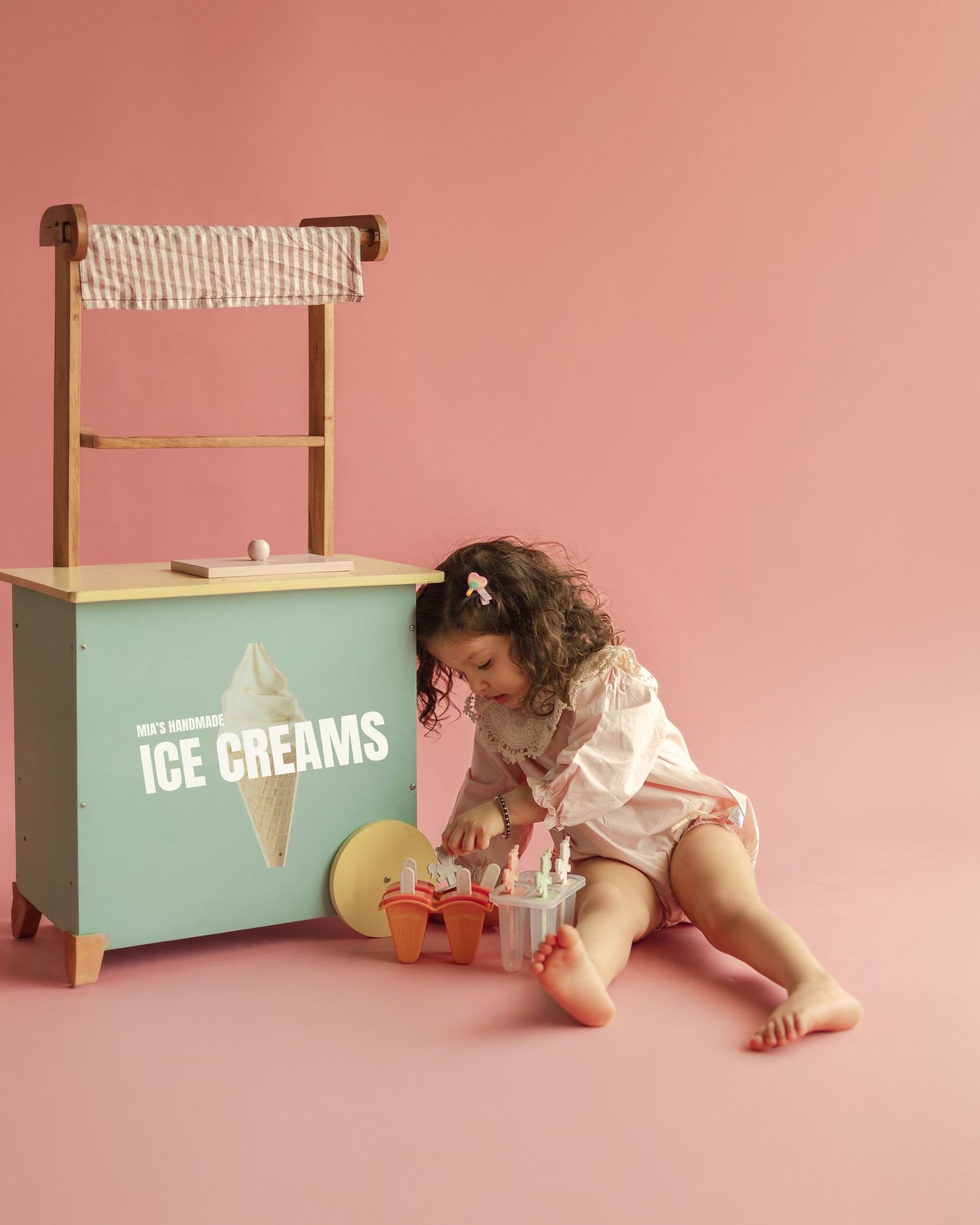 Toddler girl playing with wooden ice cream toy stand set on pink background.