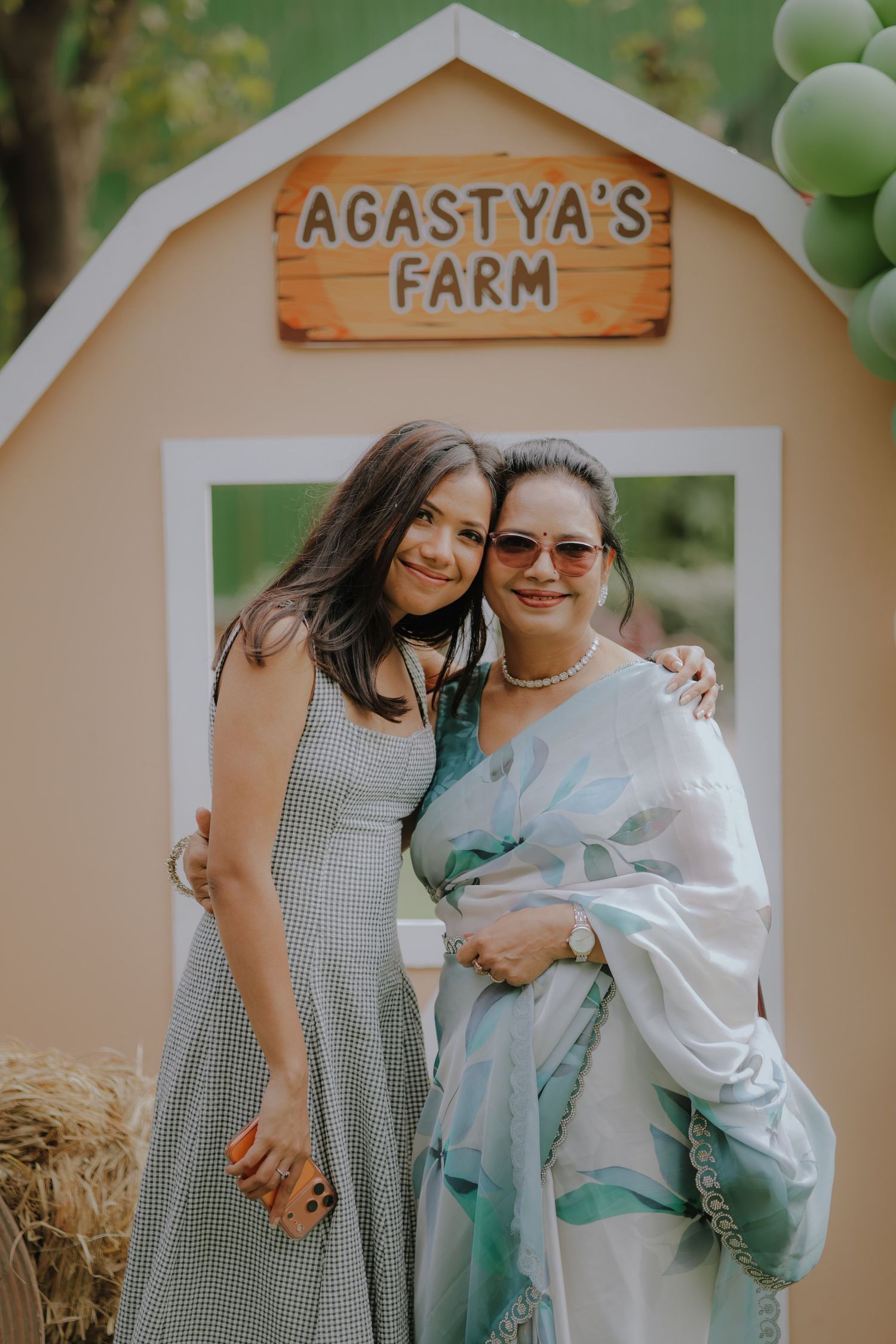 Two women smiling together at Agastya's Farm themed party backdrop with green balloon decorations.