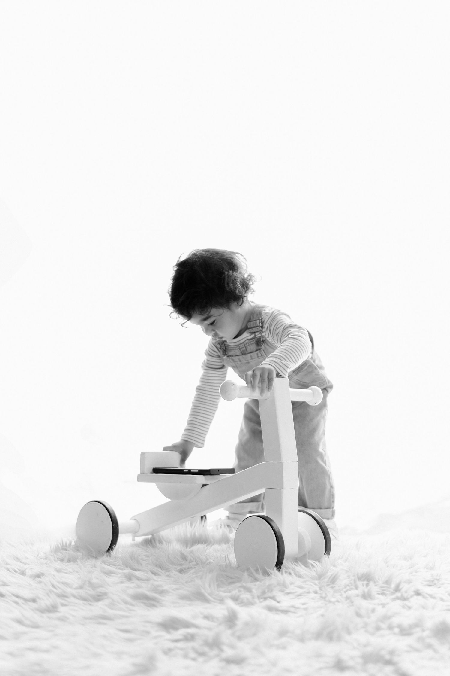Black and white photo of a toddler playing with a wooden ride-on toy on a fluffy white rug.