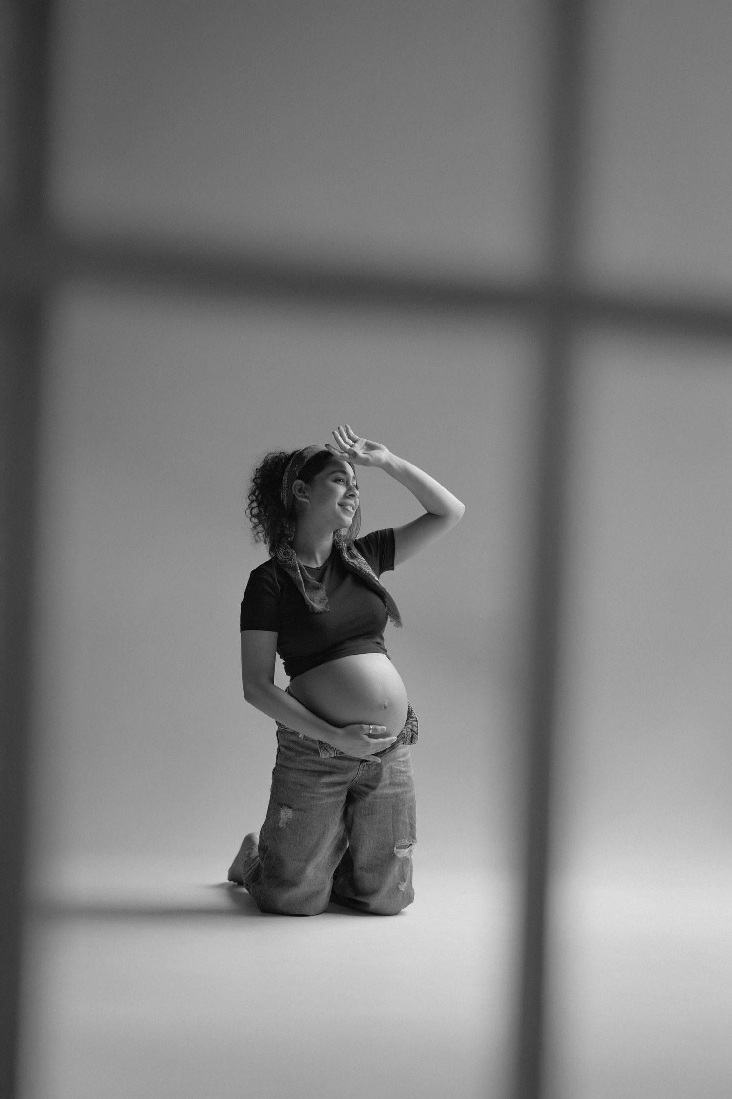 Black and white photo of a smiling pregnant woman kneeling, cradling her baby bump in a studio setting.