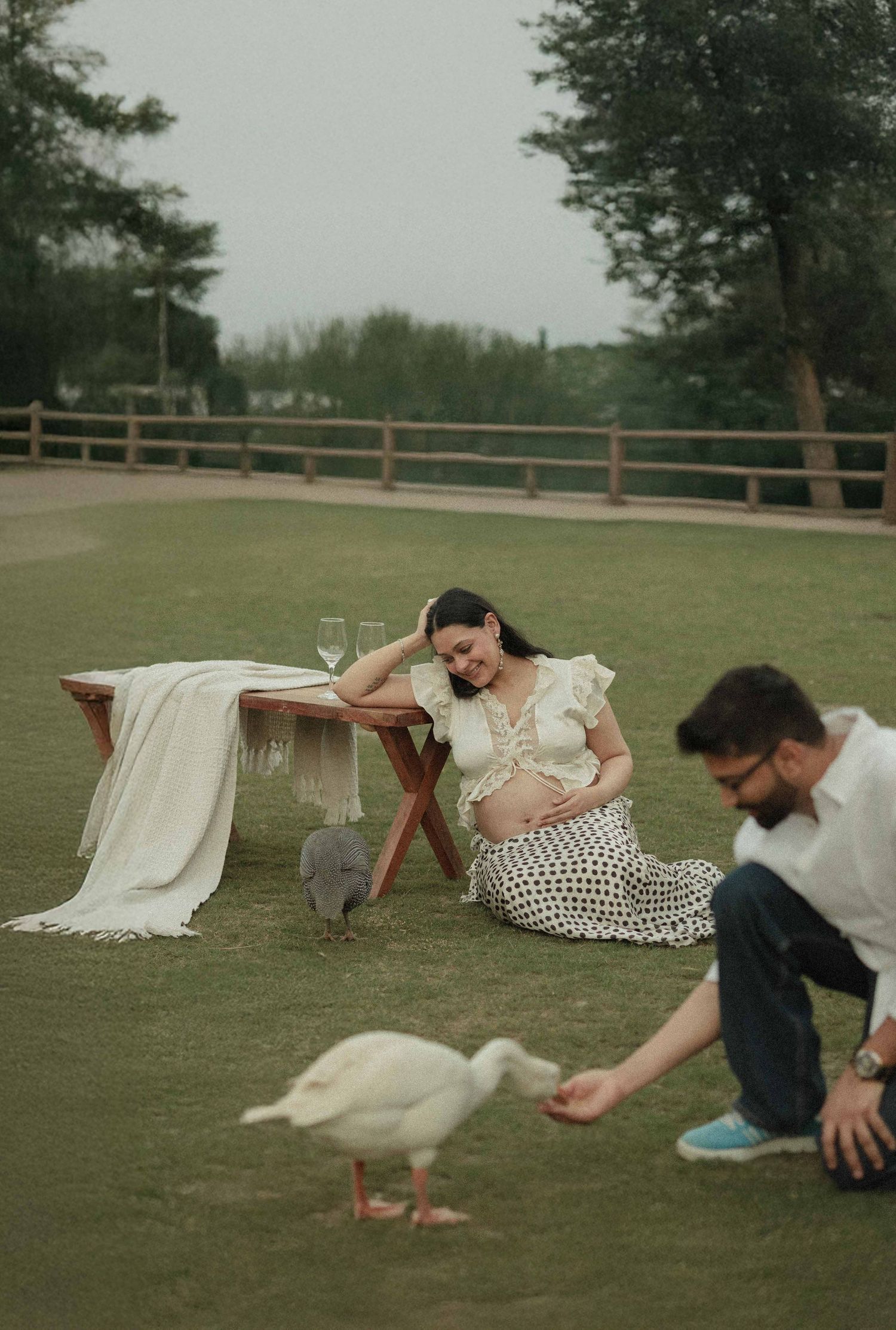 Pregnant woman sits on grass smiling, man feeds a white duck nearby, picnic table with wine glasses outdoors.
