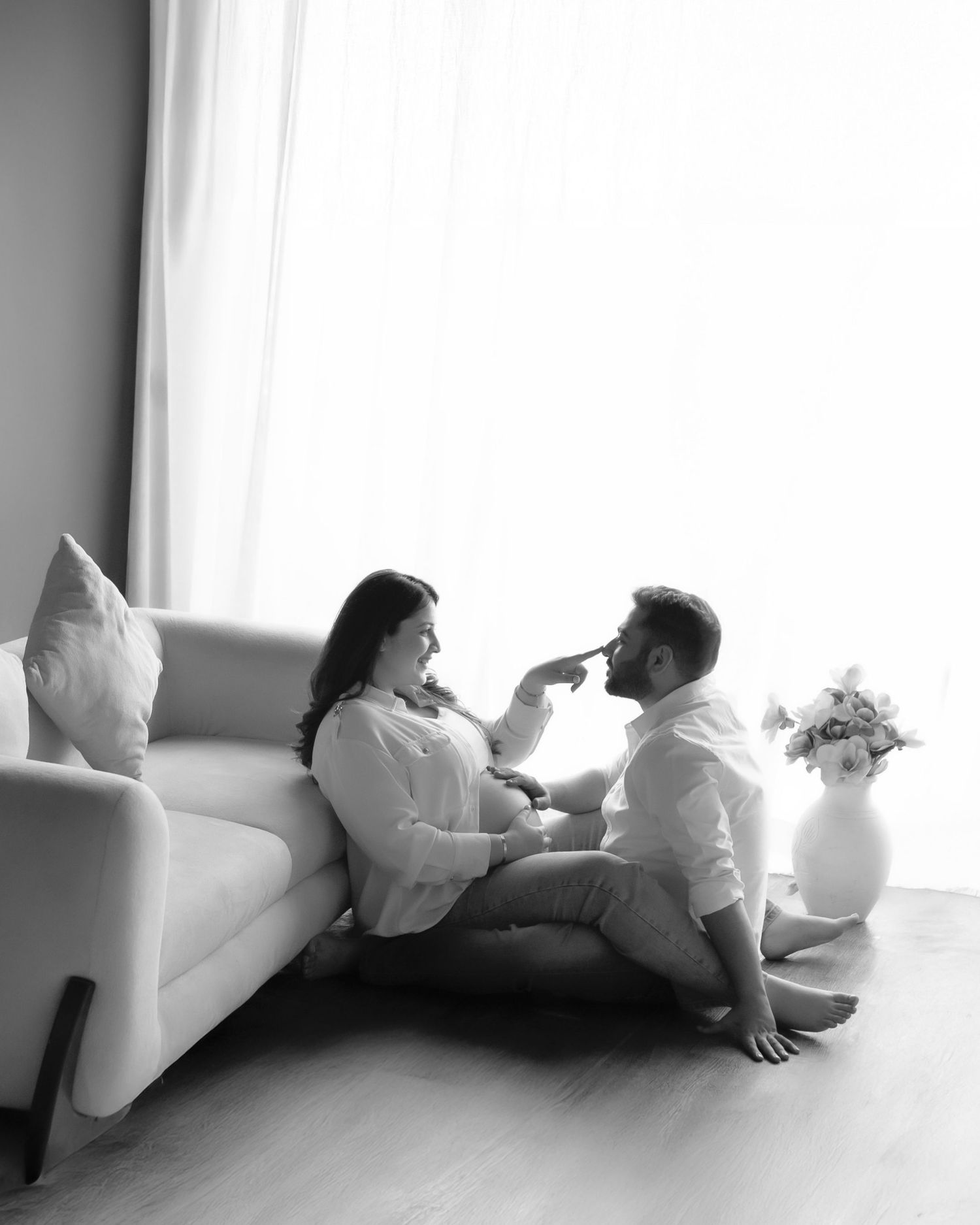Black and white photo of a romantic couple sitting on the floor by a sofa, sharing a tender moment near a window.