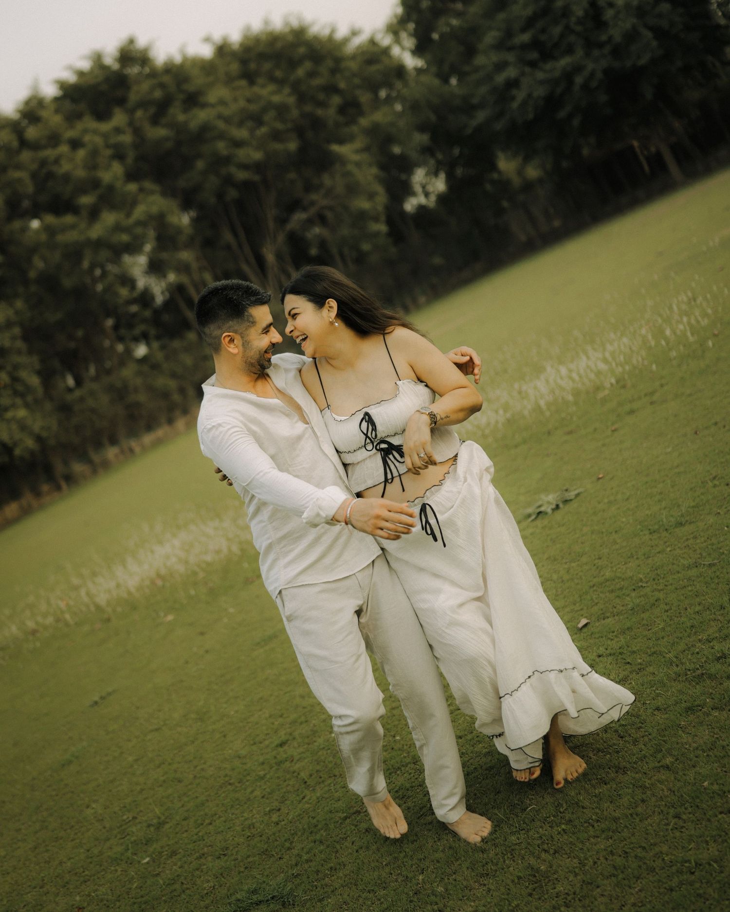 Happy couple in matching white outfits embracing and dancing barefoot on a lush green lawn.