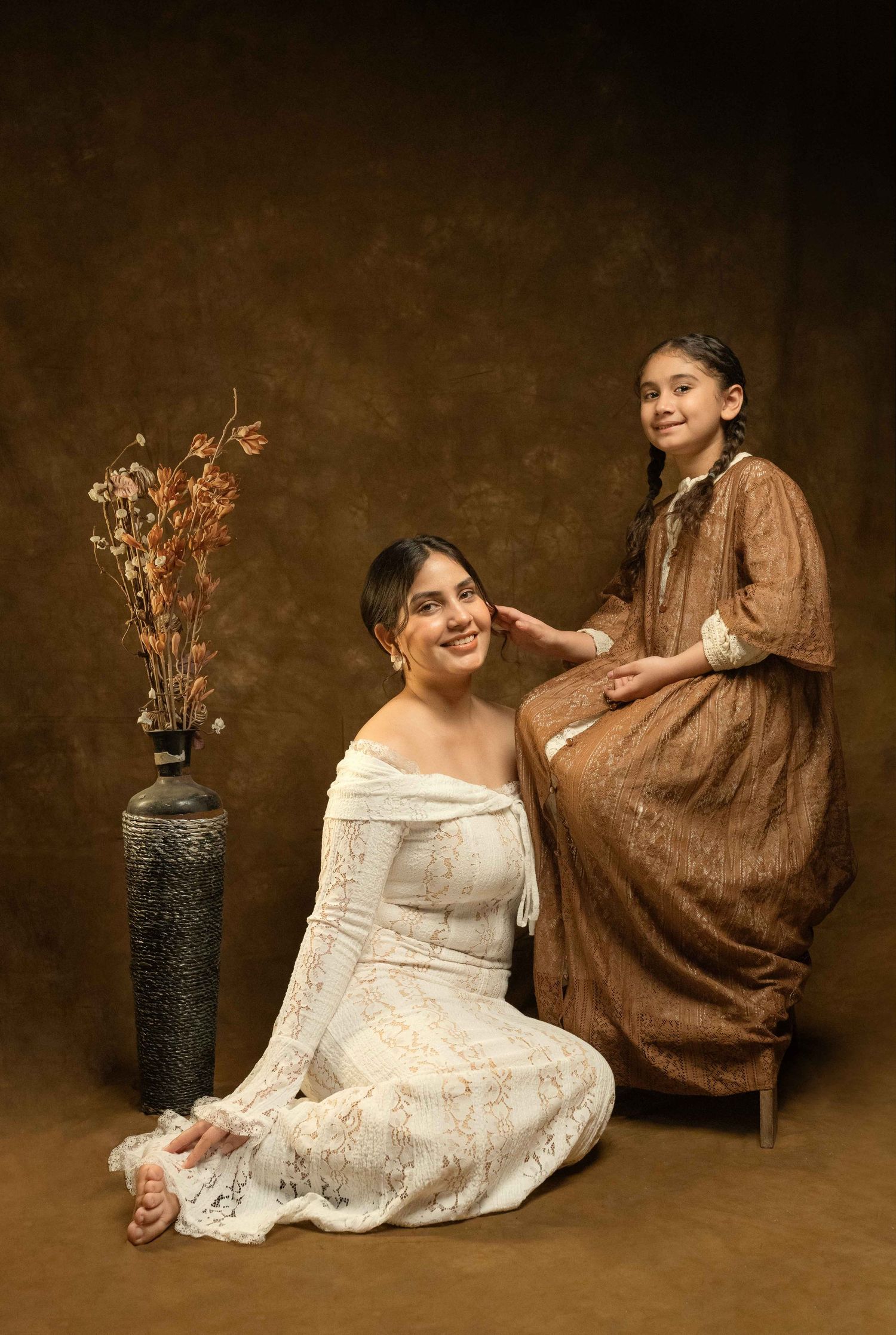 Mother in white lace dress seated with daughter in brown dress in studio portrait.