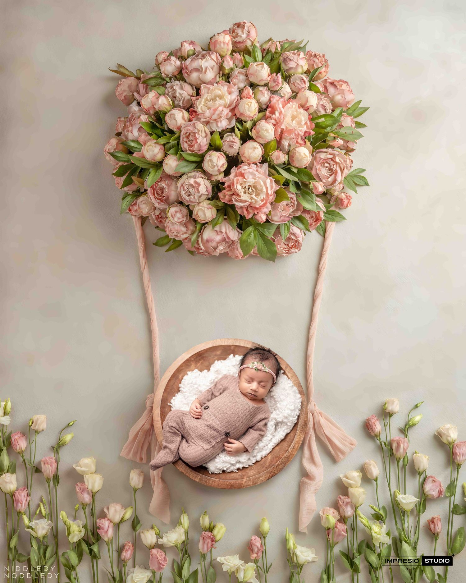 Newborn baby sleeping in wooden bowl under floral hot air balloon with pink peonies and roses in soft studio portrait.