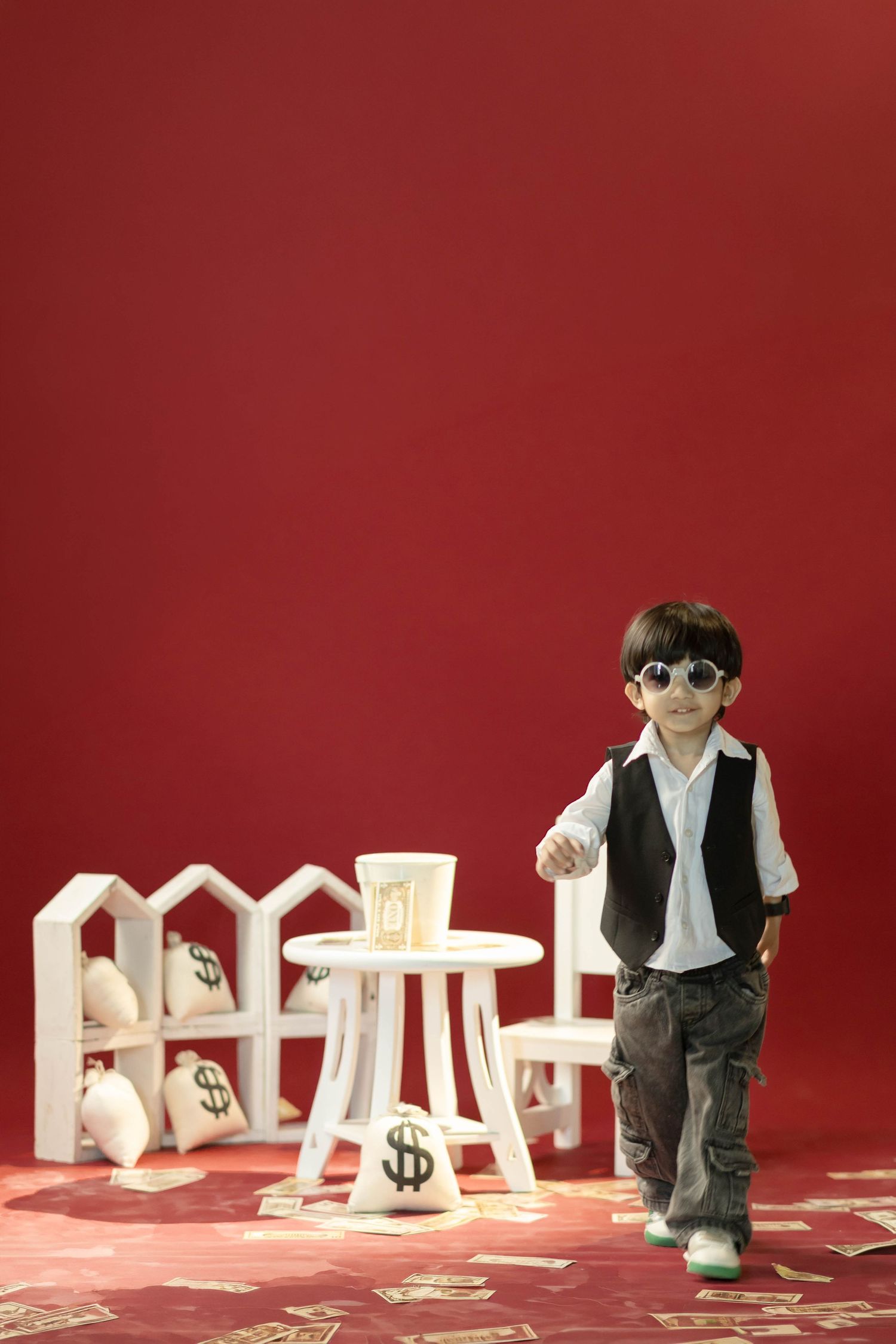 Young boy in sunglasses and vest poses confidently with money bags and dollar bills on a red background.