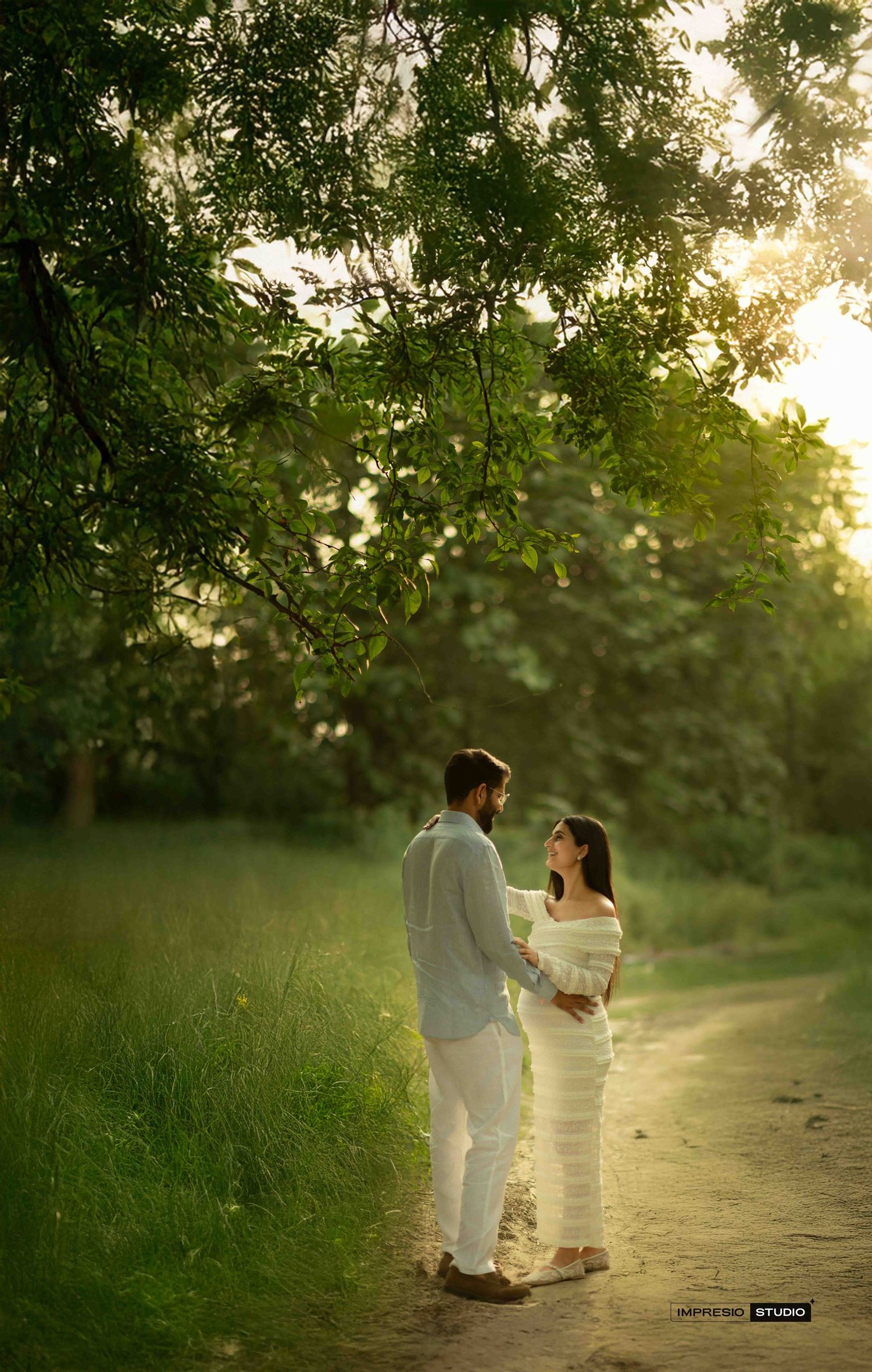 A couple in white outfits holds hands on a sunlit garden path surrounded by lush green trees during golden hour.
