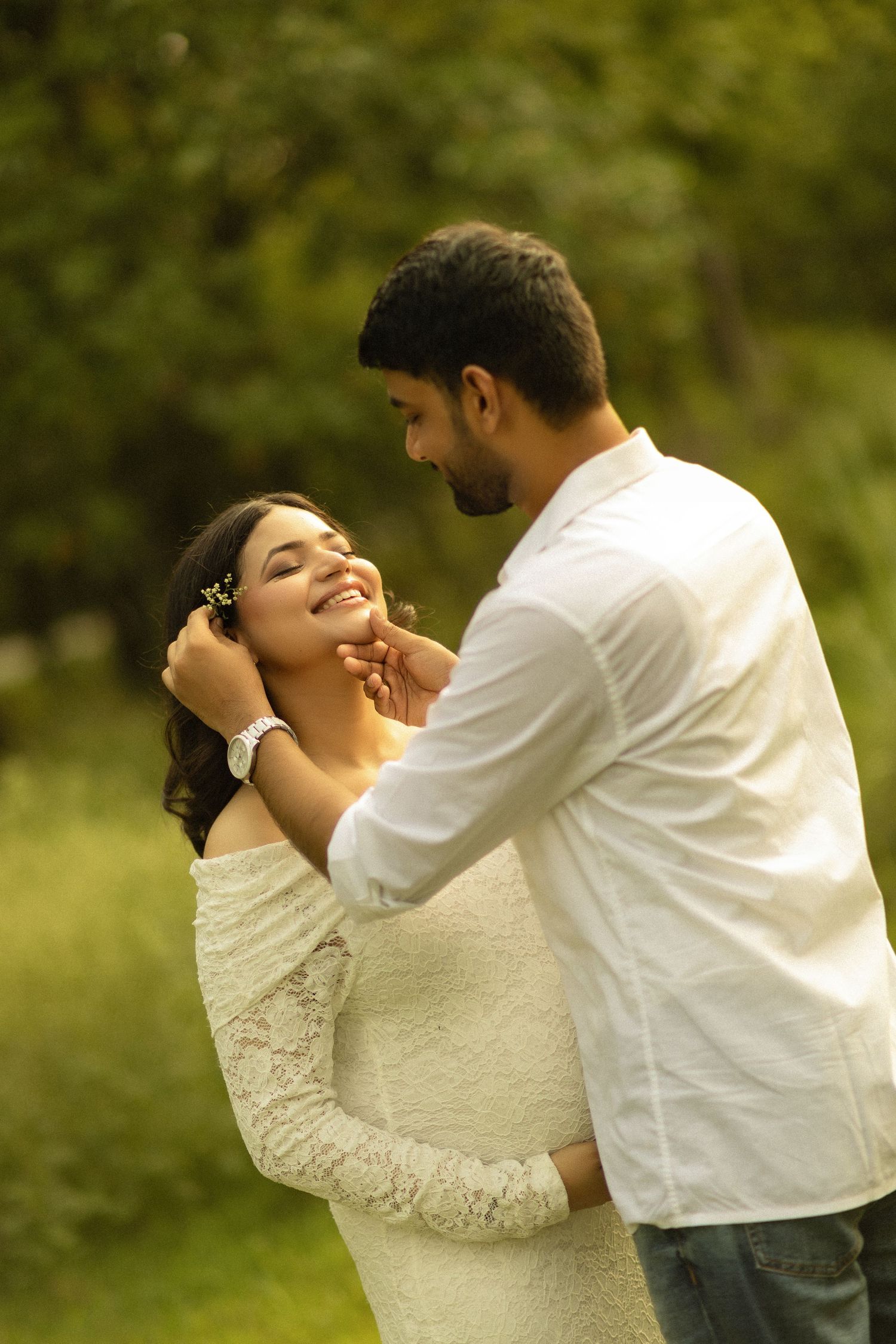 A romantic moment between a couple dressed in white embracing in a sunlit outdoor maternity photoshoot setting with green foliage.