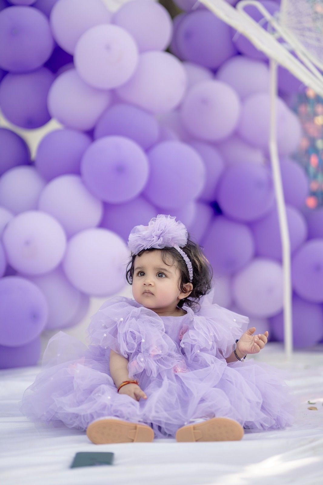 A small child in a lavender tulle dress poses in front of a purple balloon wall backdrop for a photoshoot.