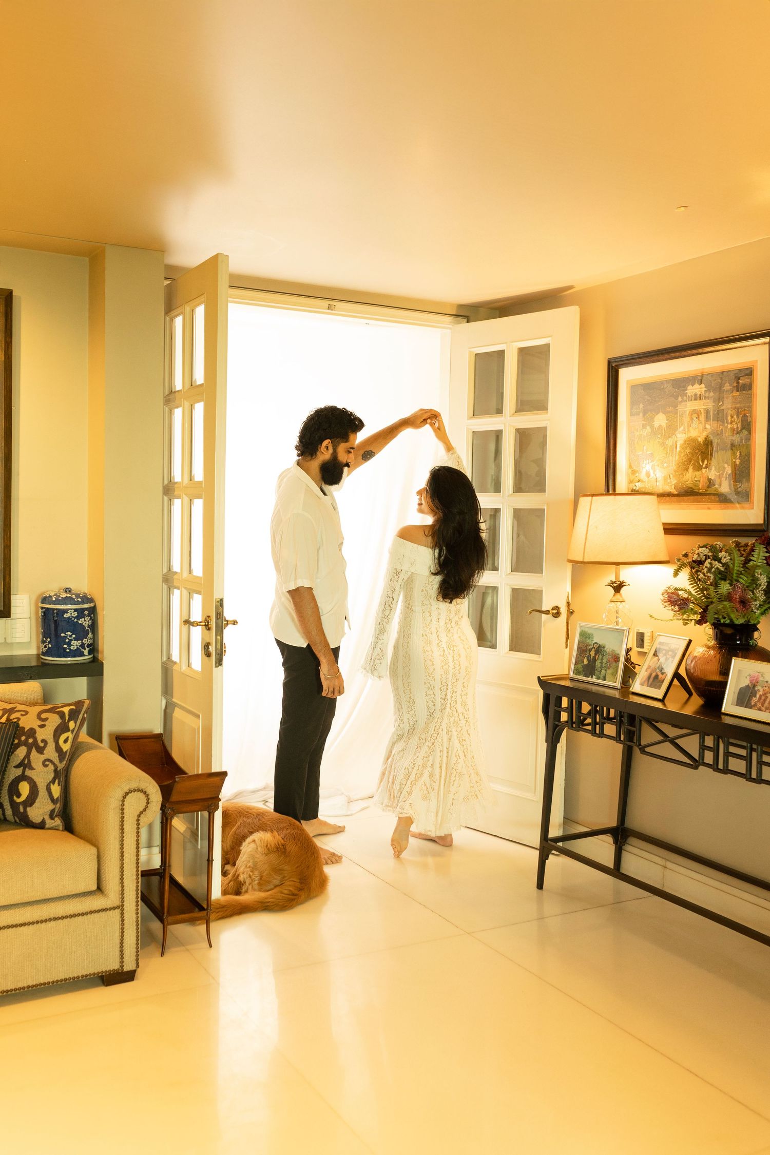 A couple dancing in a warmly lit living room with French doors while a ginger cat relaxes on a beige sofa for indoor maternity photography.