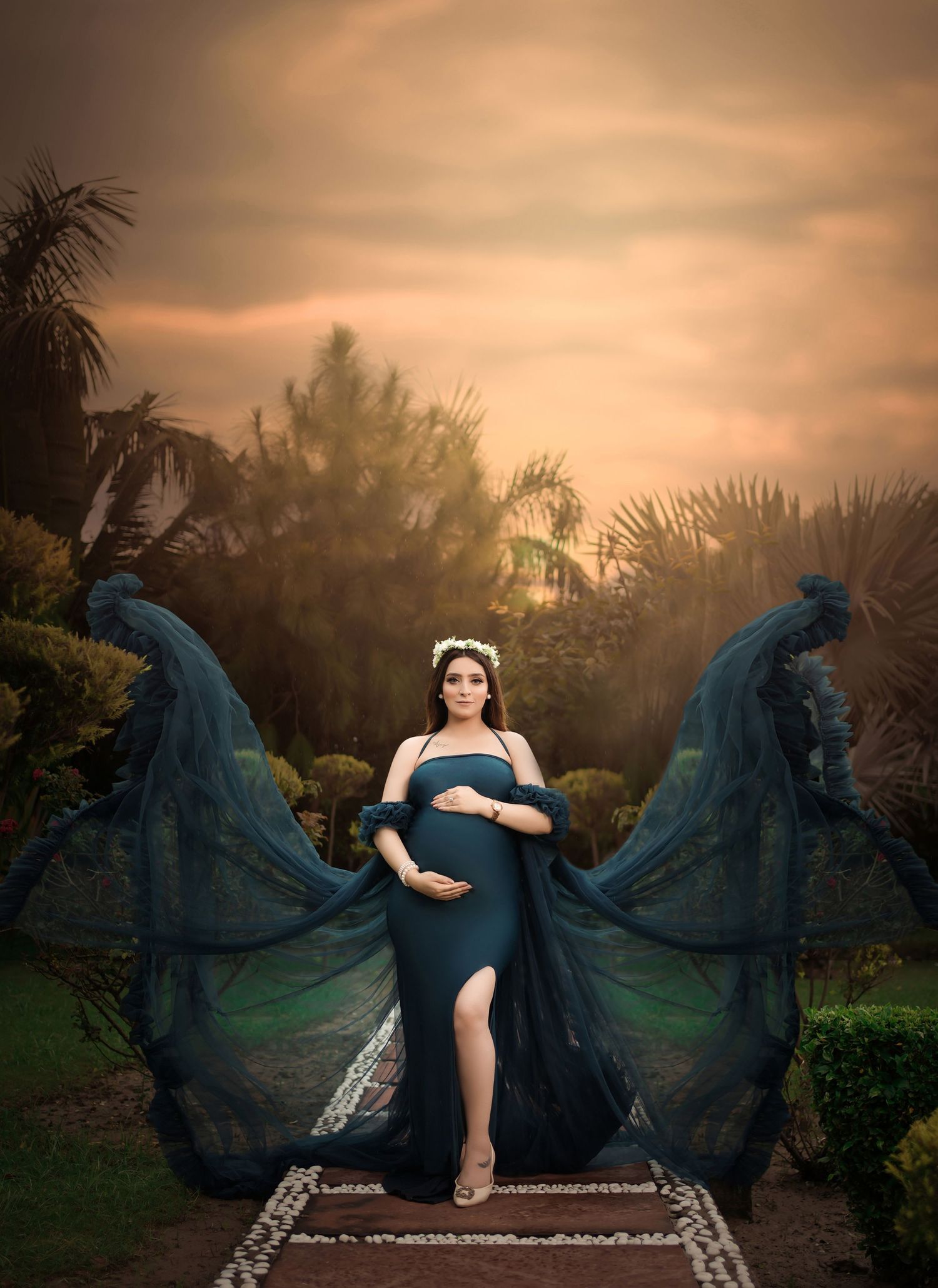 Dramatic maternity photo shoot featuring flowing teal gown against sunset backdrop with dreamy lighting.