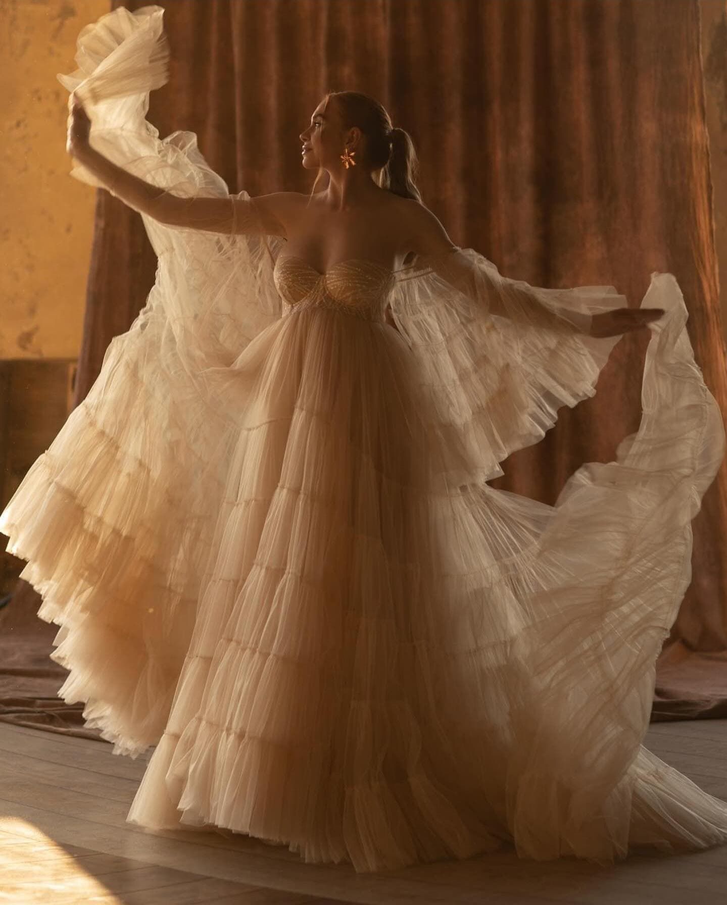Romantic cream tulle gown photographed in dramatic backlit setting with flowing fabric movement.