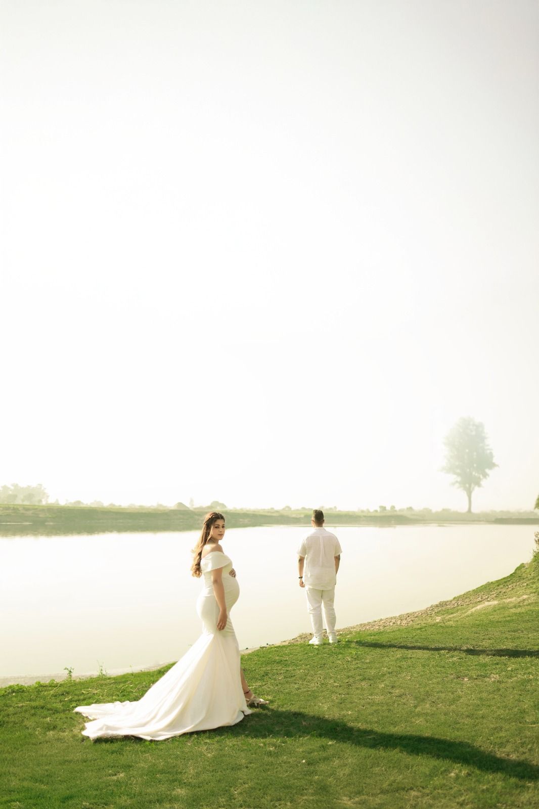 Bride and groom walking along grassy lakeside path in ethereal lighting.