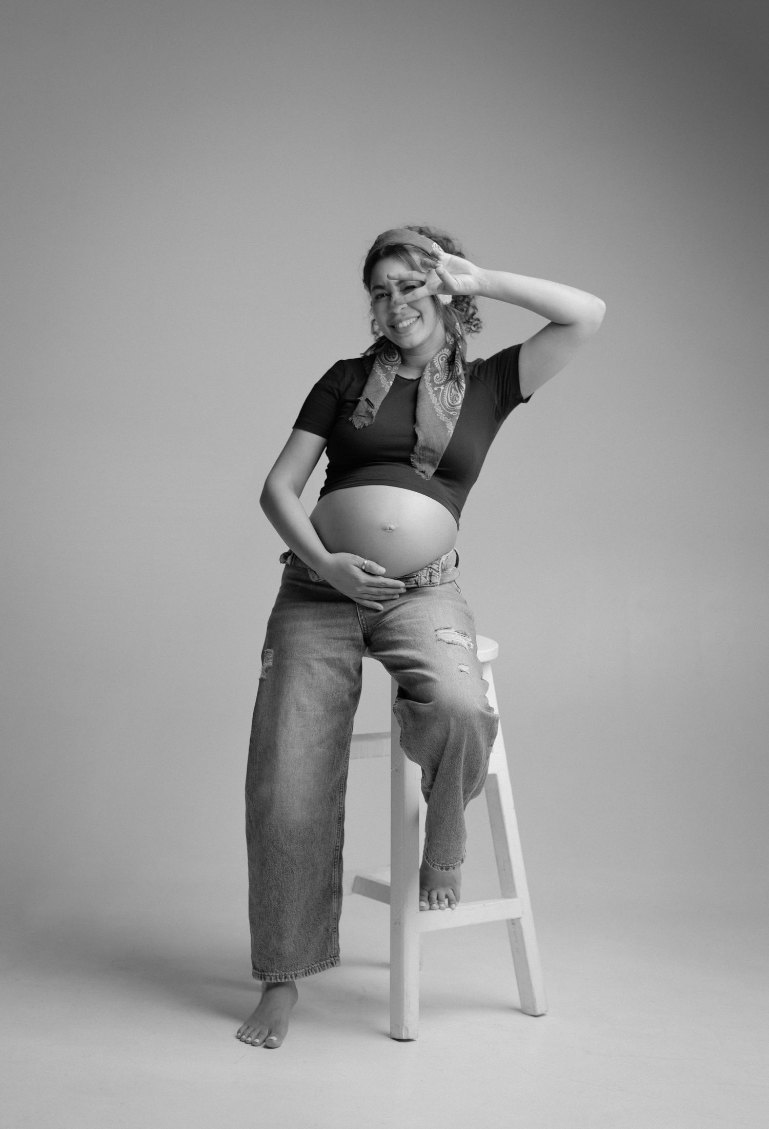 Black and white maternity photo of smiling pregnant woman in crop top and jeans, posing on stool with peace sign.