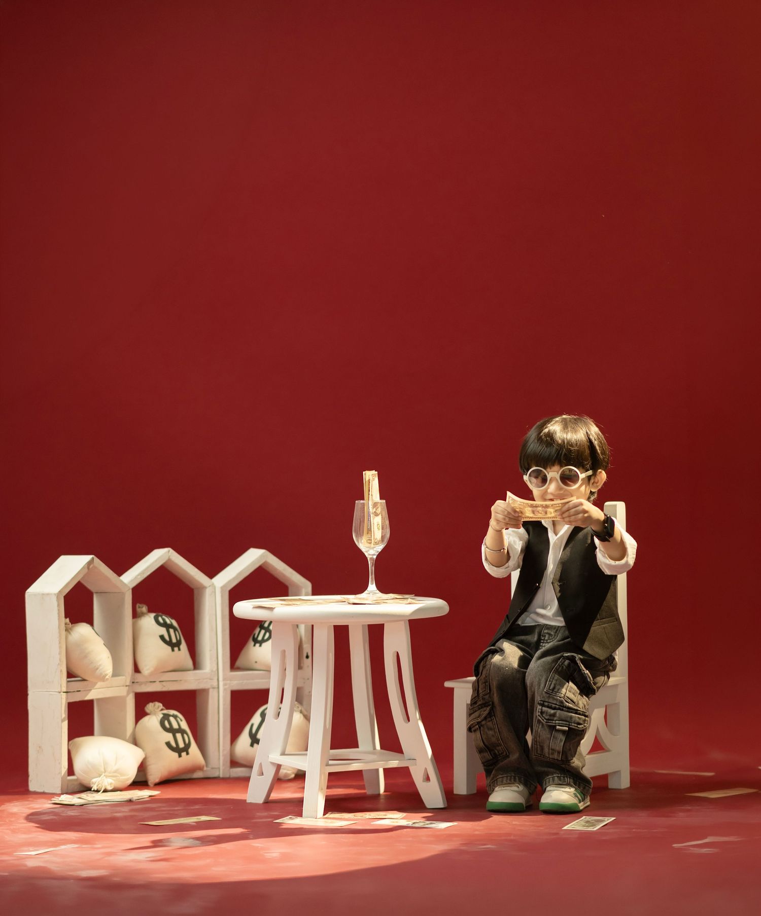 Young child in sunglasses sits on white chair with money bags and wine glass on red background.