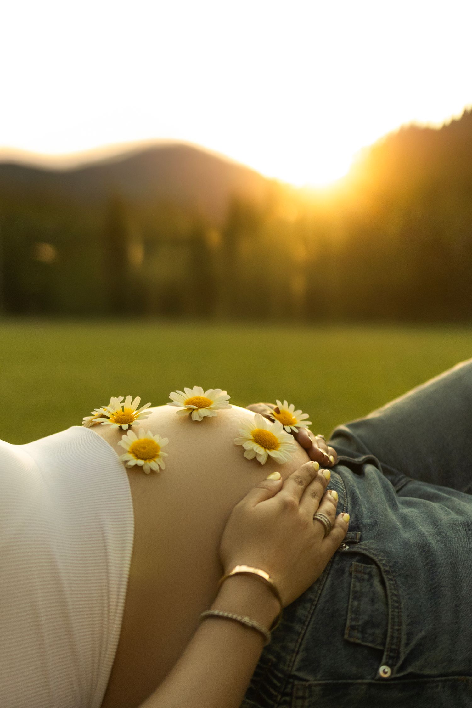 Pregnant woman with daisies on belly at golden sunset in a meadow with mountains in background.