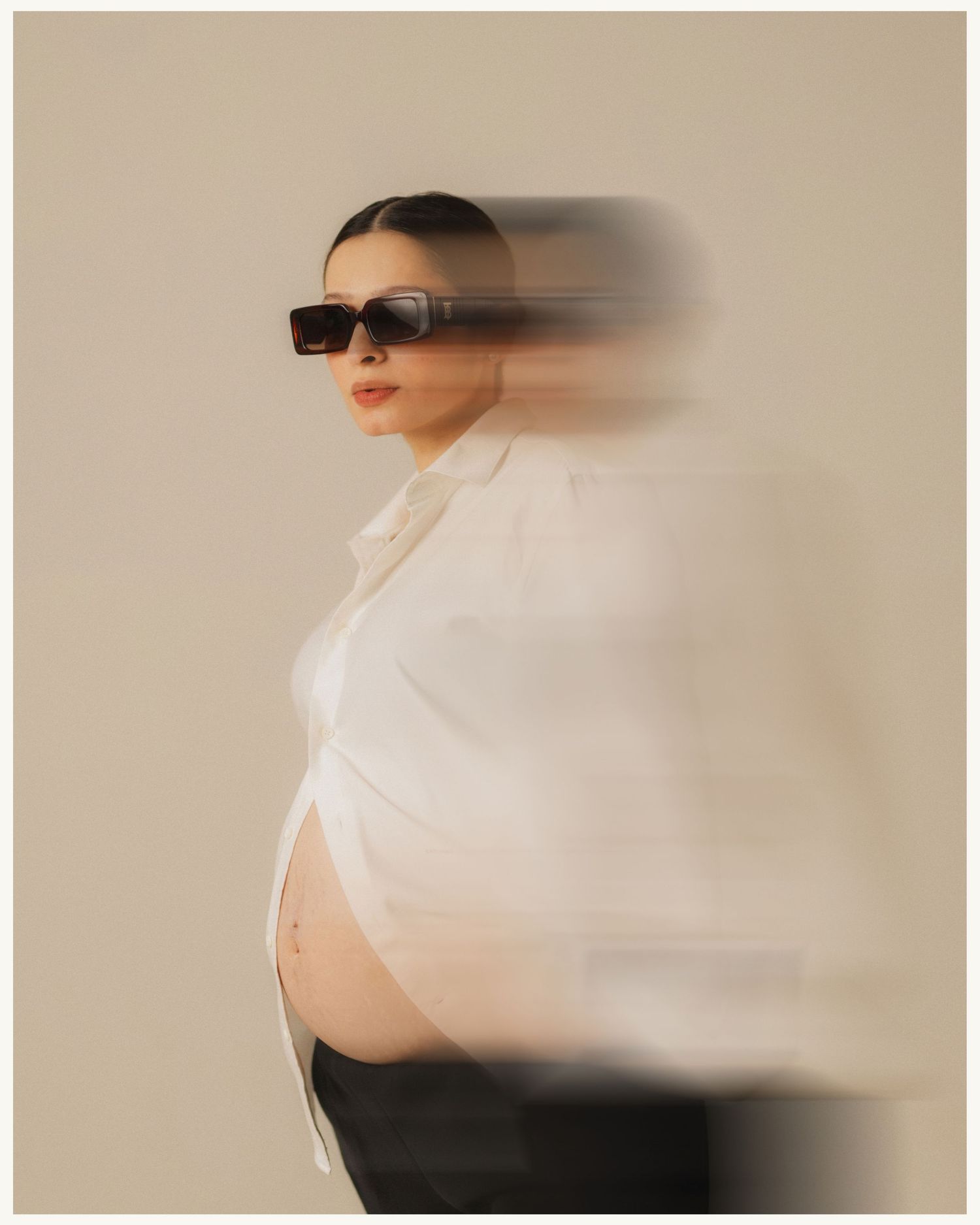 Pregnant woman in white shirt and black sunglasses poses with artistic motion blur effect against neutral background.