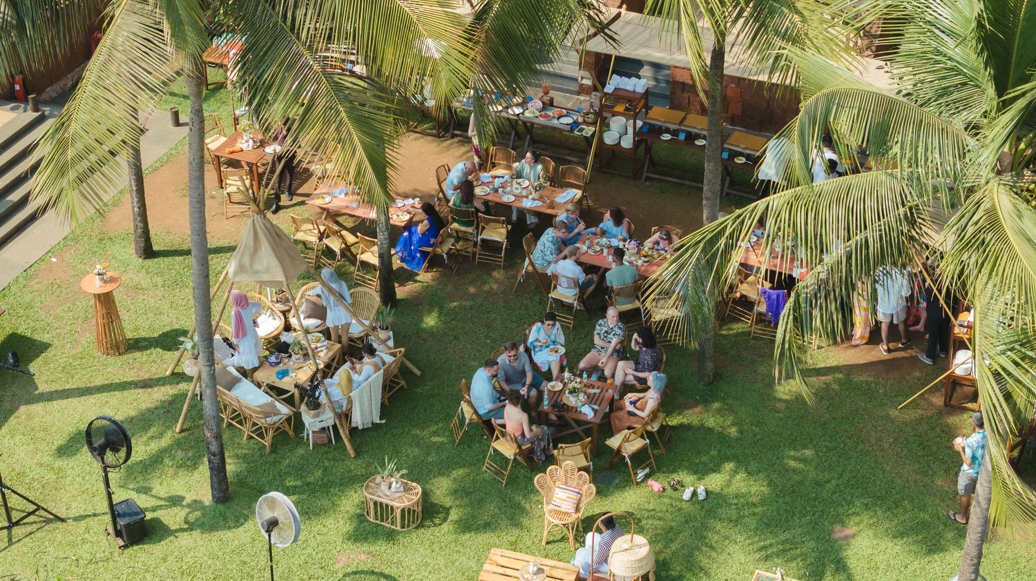 Aerial view of guests dining outdoors at a tropical restaurant surrounded by palm trees on lush green grass.