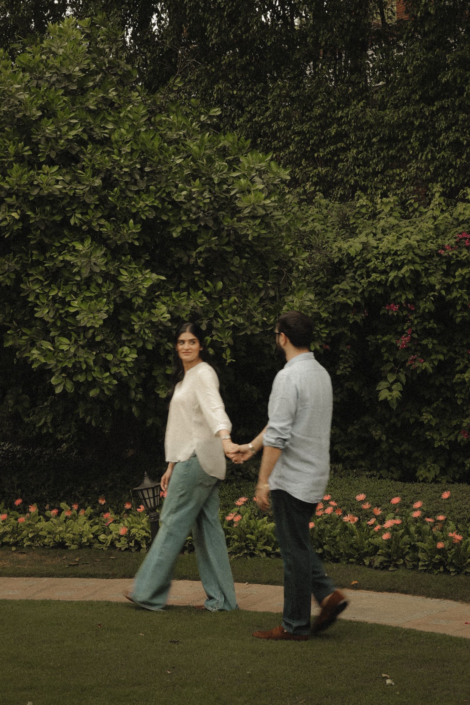 Couple holding hands walking in a lush garden with green trees and pink flowers in the background.