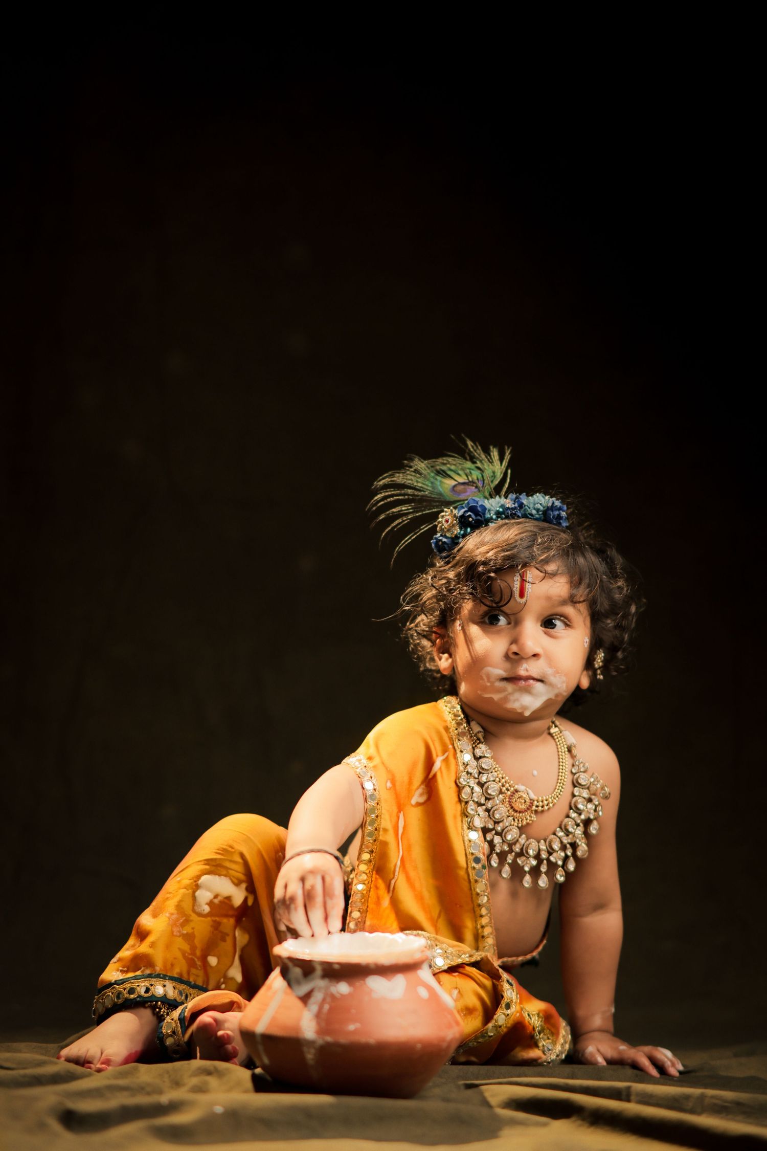 Young child dressed as Lord Krishna in yellow outfit, peacock feather crown, and gold jewelry beside a clay pot.