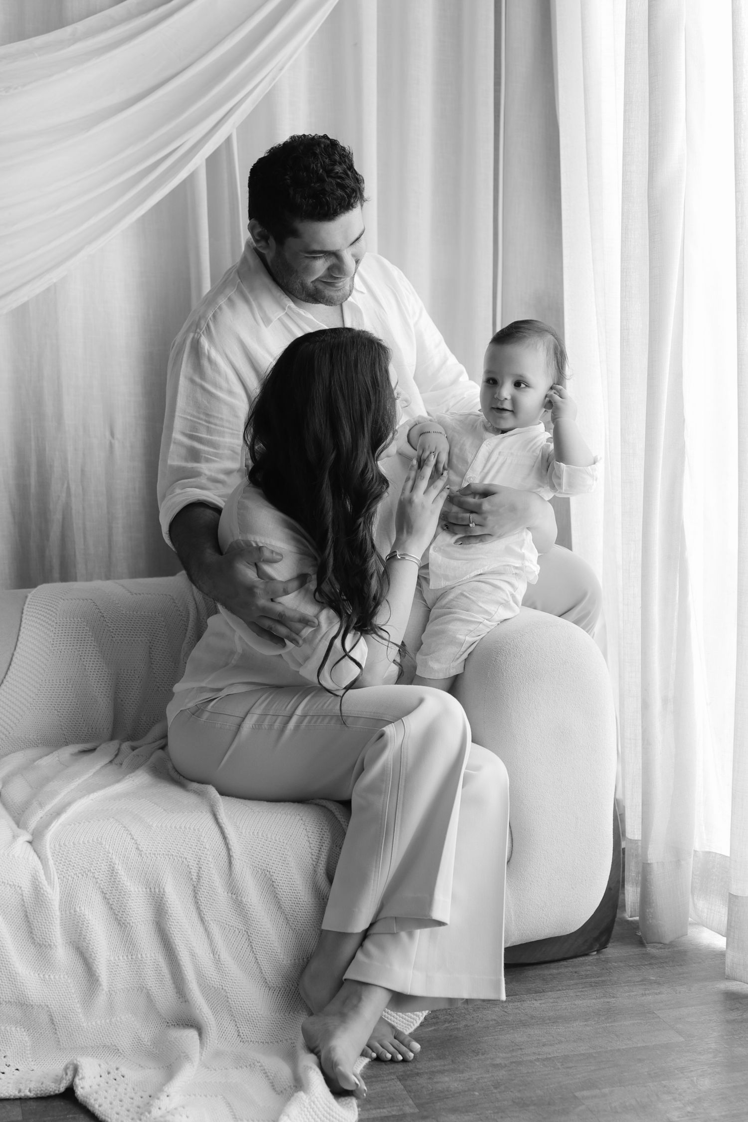 Black and white family portrait of parents and baby sitting on sofa near curtained window.