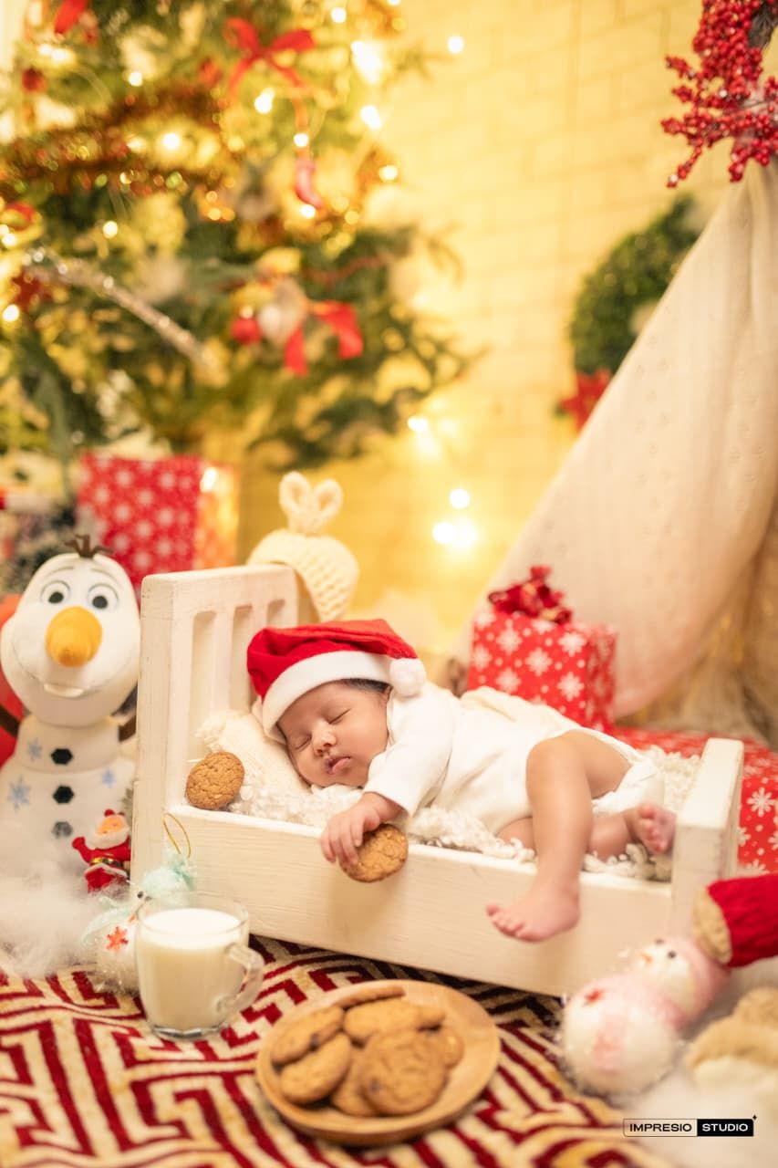 Newborn baby sleeping in wooden crate wearing Santa hat, surrounded by Christmas tree, cookies, milk, and Olaf snowman toy.