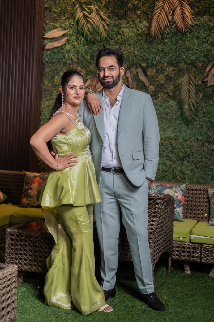 Elegant couple posing at a party, woman in green outfit and man in light gray suit against a tropical green wall backdrop.