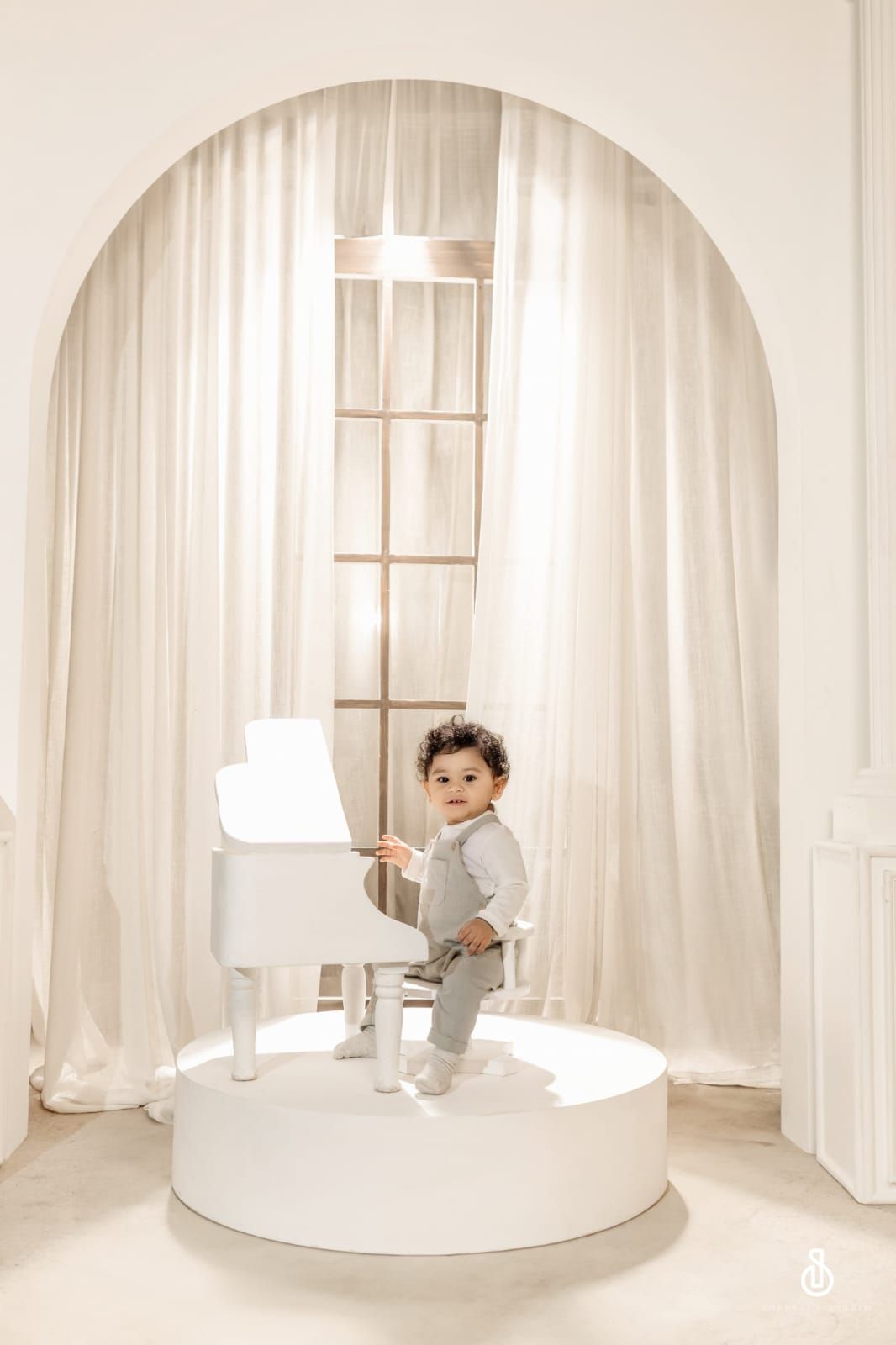 Toddler in gray outfit playing a white mini grand piano on a circular platform in an elegant white studio.