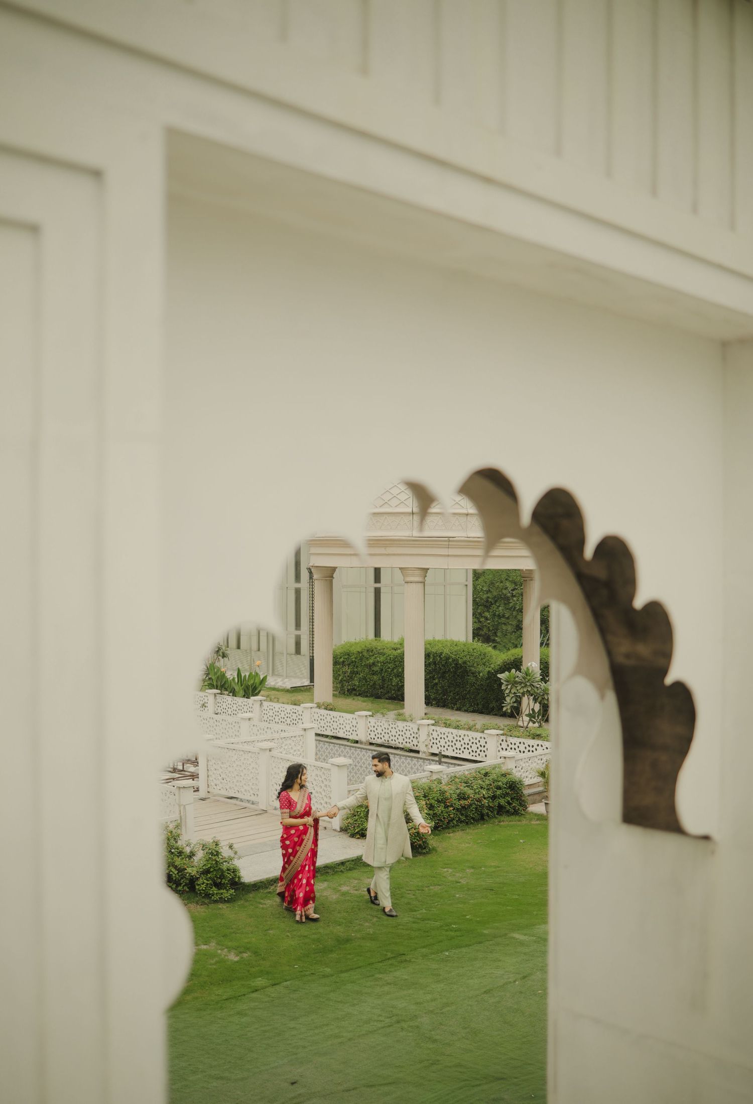 Two people in elegant attire walk together through a lush garden viewed through an ornate archway opening.