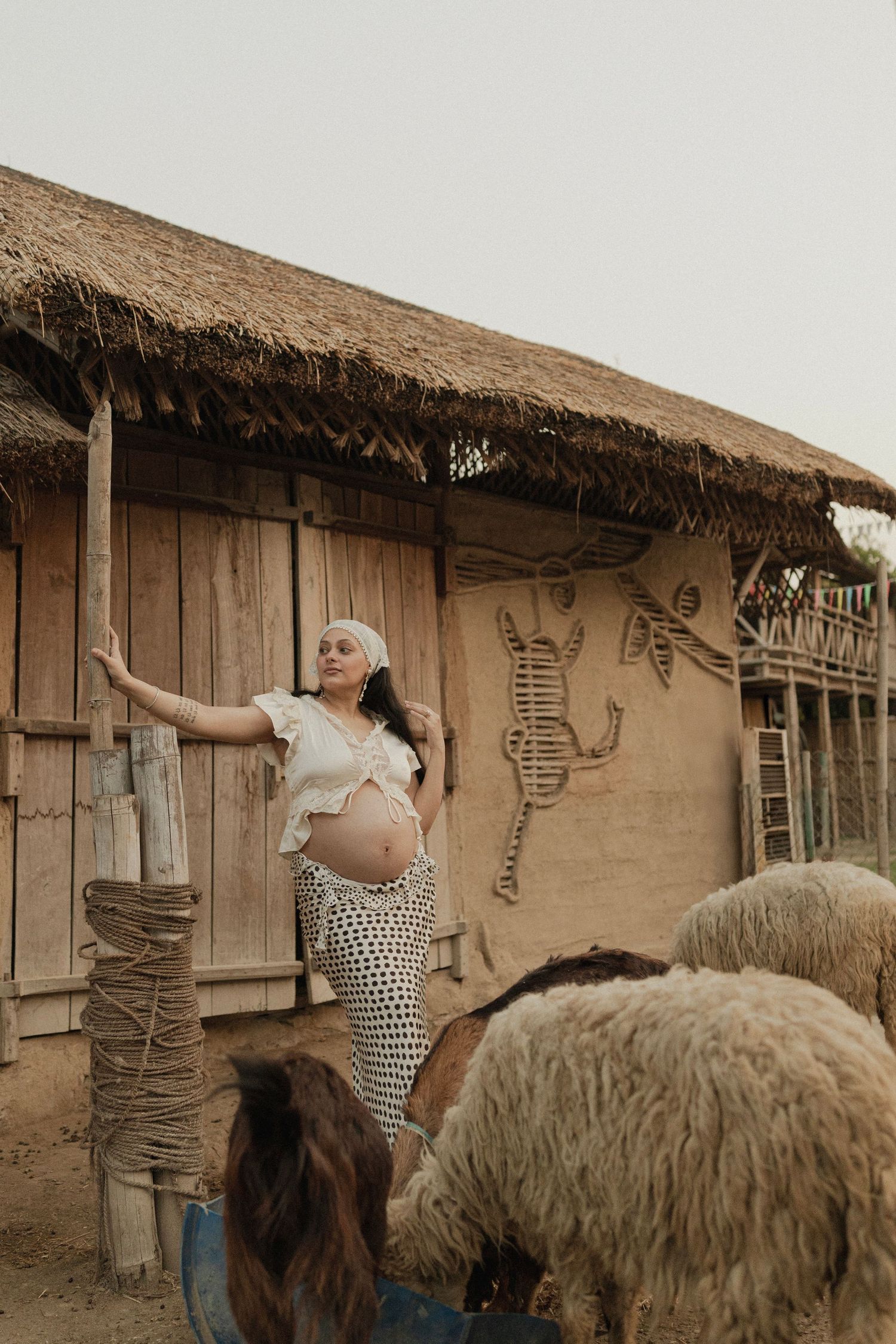 Pregnant woman in white top and polka dot skirt poses by rustic mud hut with thatched roof and sheep nearby.