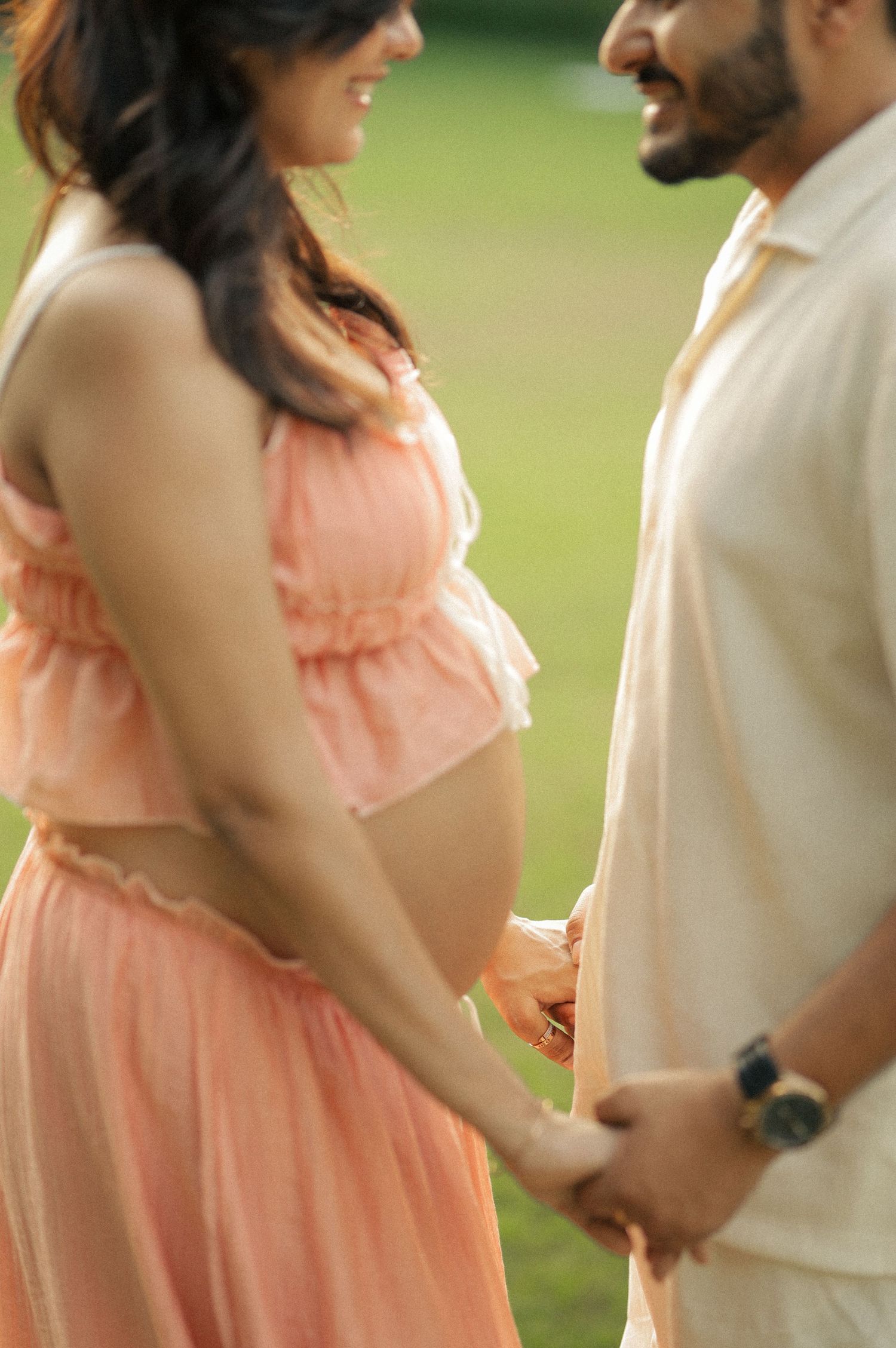 Pregnant couple holding hands outdoors, woman in peach maternity dress showing baby bump, man in cream shirt smiling.