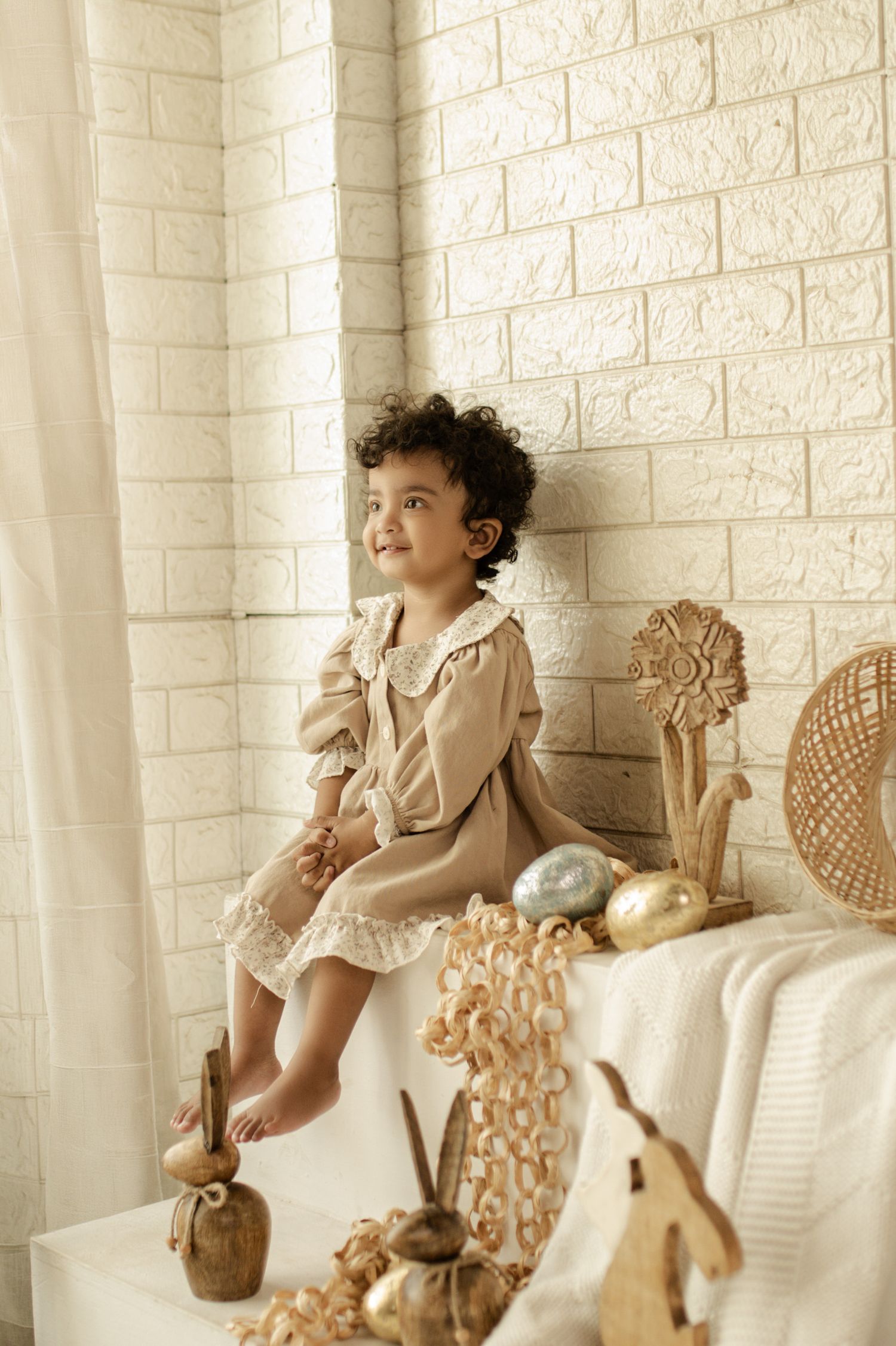 Young girl in beige lace dress sitting against white brick wall with wicker decorations and golden Easter eggs.