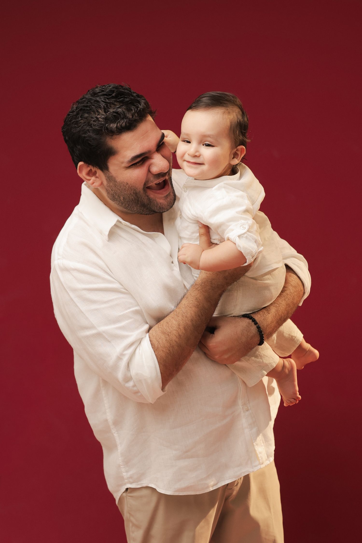 Happy father in white shirt holding smiling baby against red background.