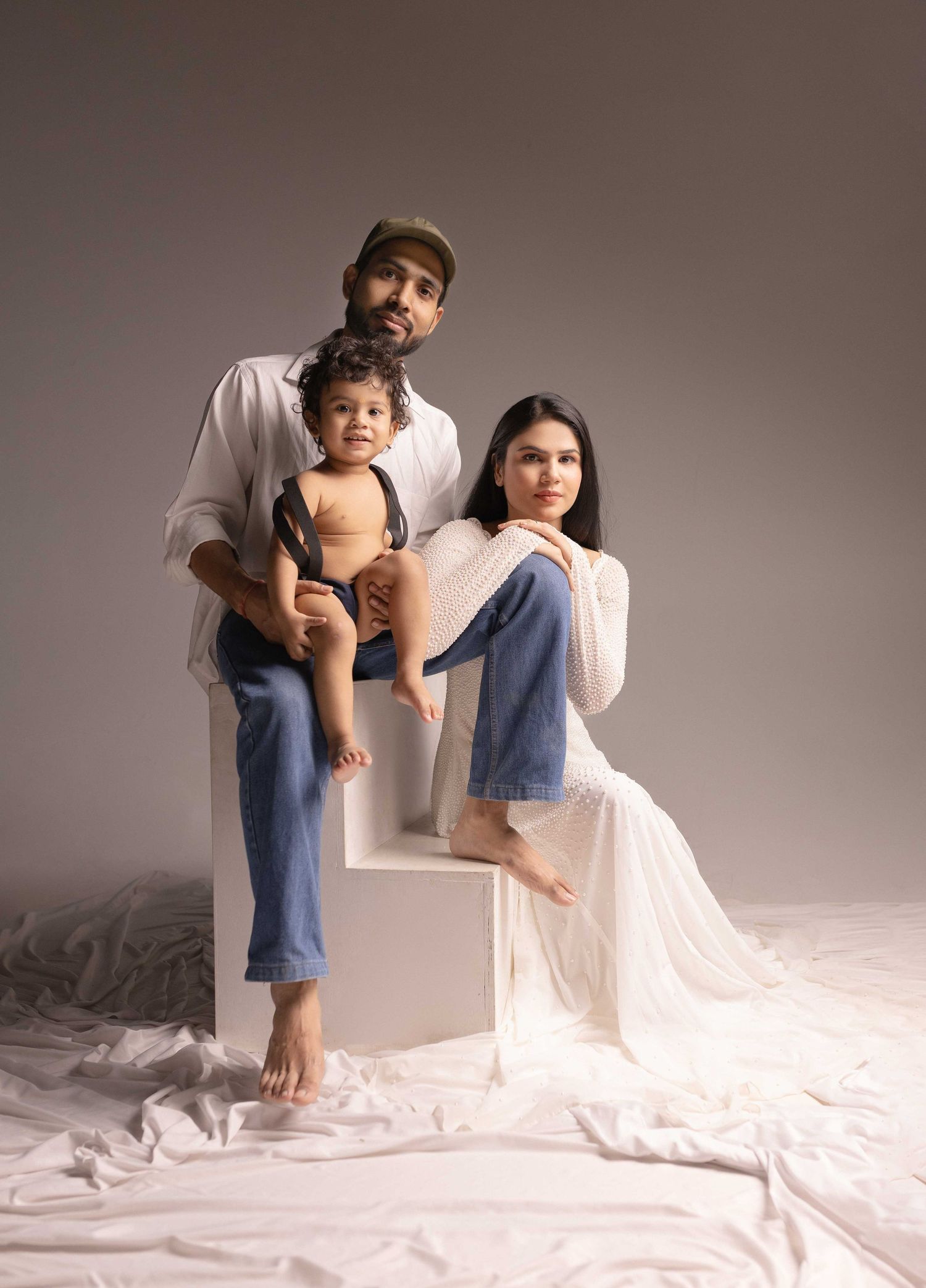 A family of three poses in a studio, with a man holding a baby and a woman in a white dress seated beside them.