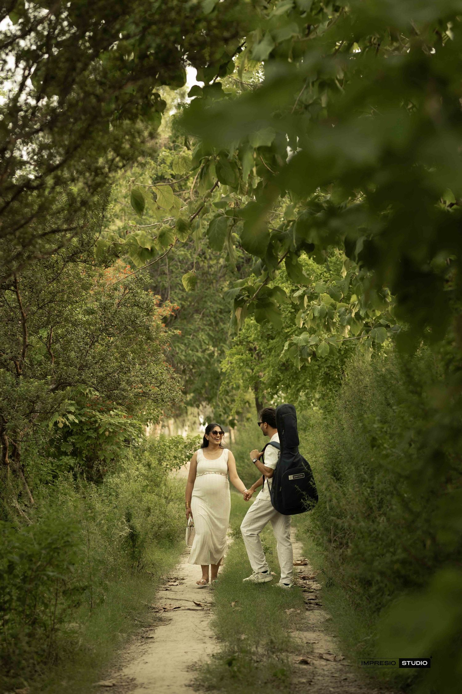 Pregnant woman in white dress holds hands with partner on a lush green forest path during maternity photoshoot.