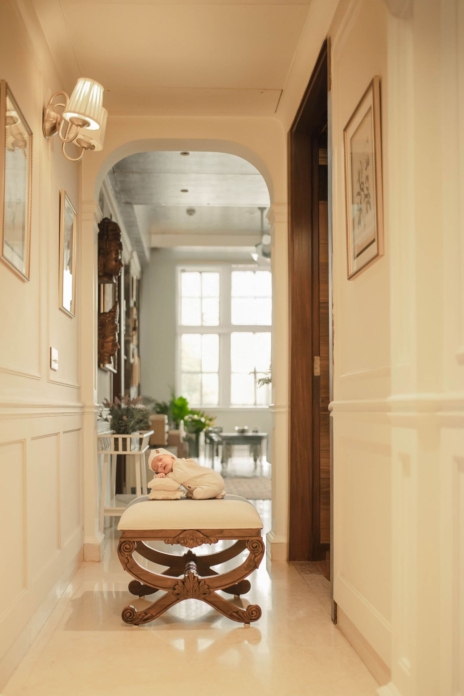 Elegant hallway with arched doorway, ornate bench holding a sleeping baby, wall sconces, and framed art in a classic home.