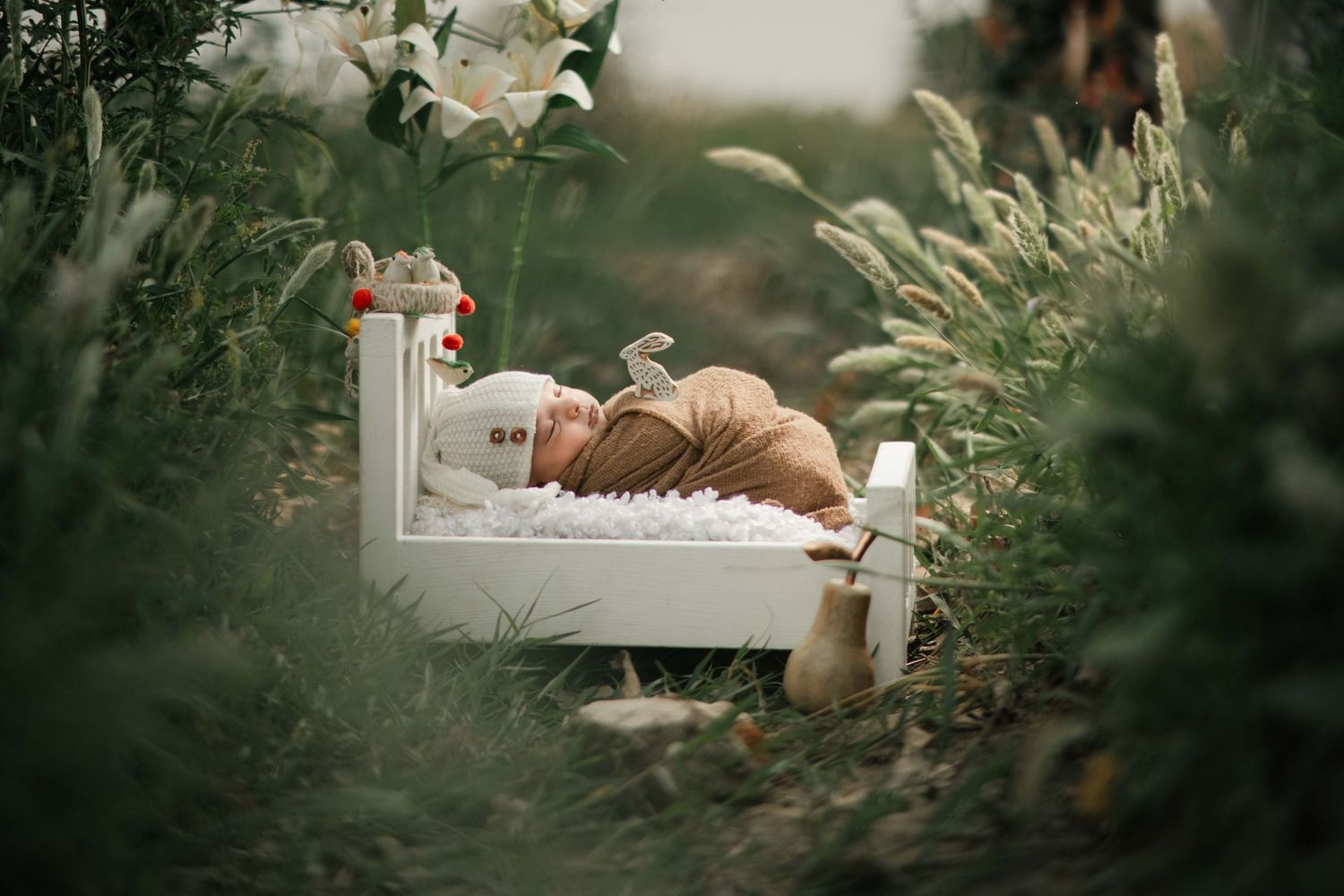 Newborn baby sleeping peacefully on a tiny white bed surrounded by lush green plants and white flowers outdoors.