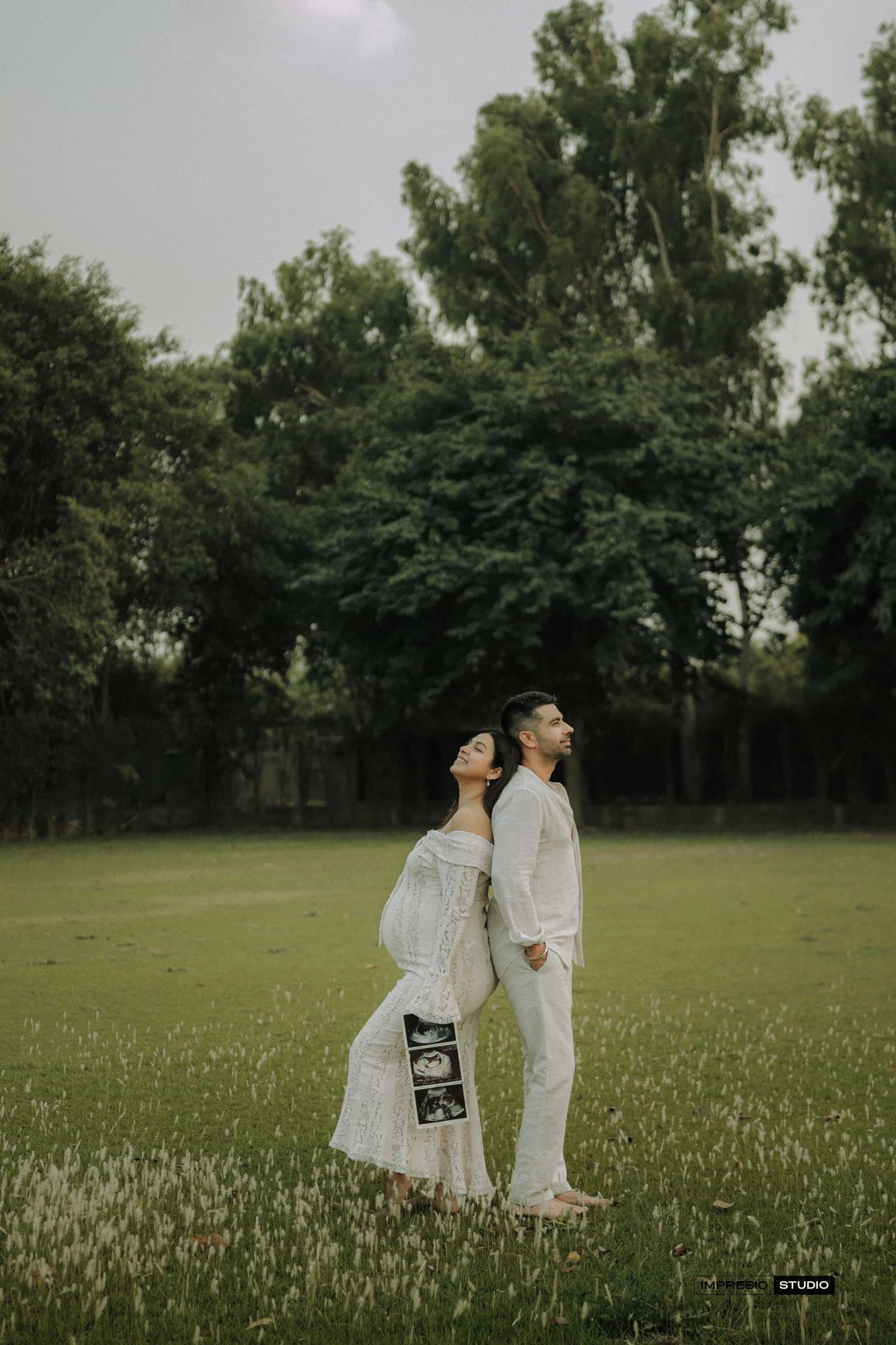 Couple in white outfits standing in green park, woman holding ultrasound scan, maternity photoshoot outdoors.