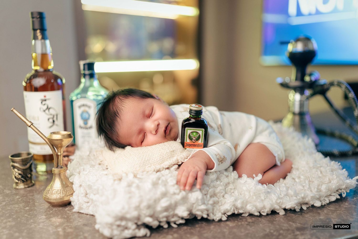 Newborn baby sleeping on fluffy white rug surrounded by liquor bottles in a creative photo shoot.