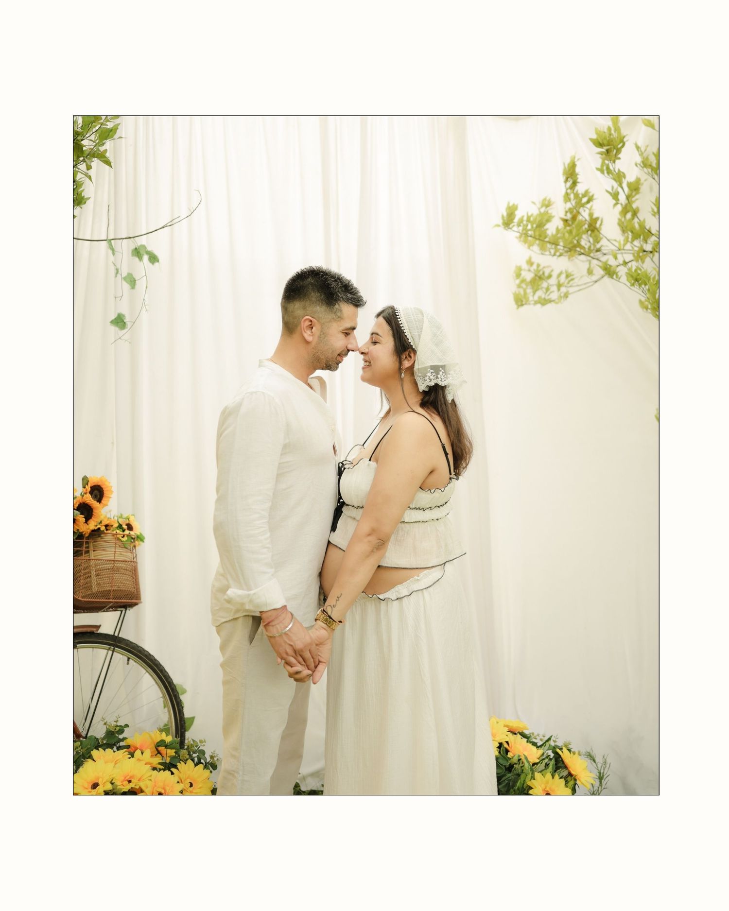 Couple in white outfits holding hands near sunflowers and bicycle, sharing a romantic moment against sheer white curtains.