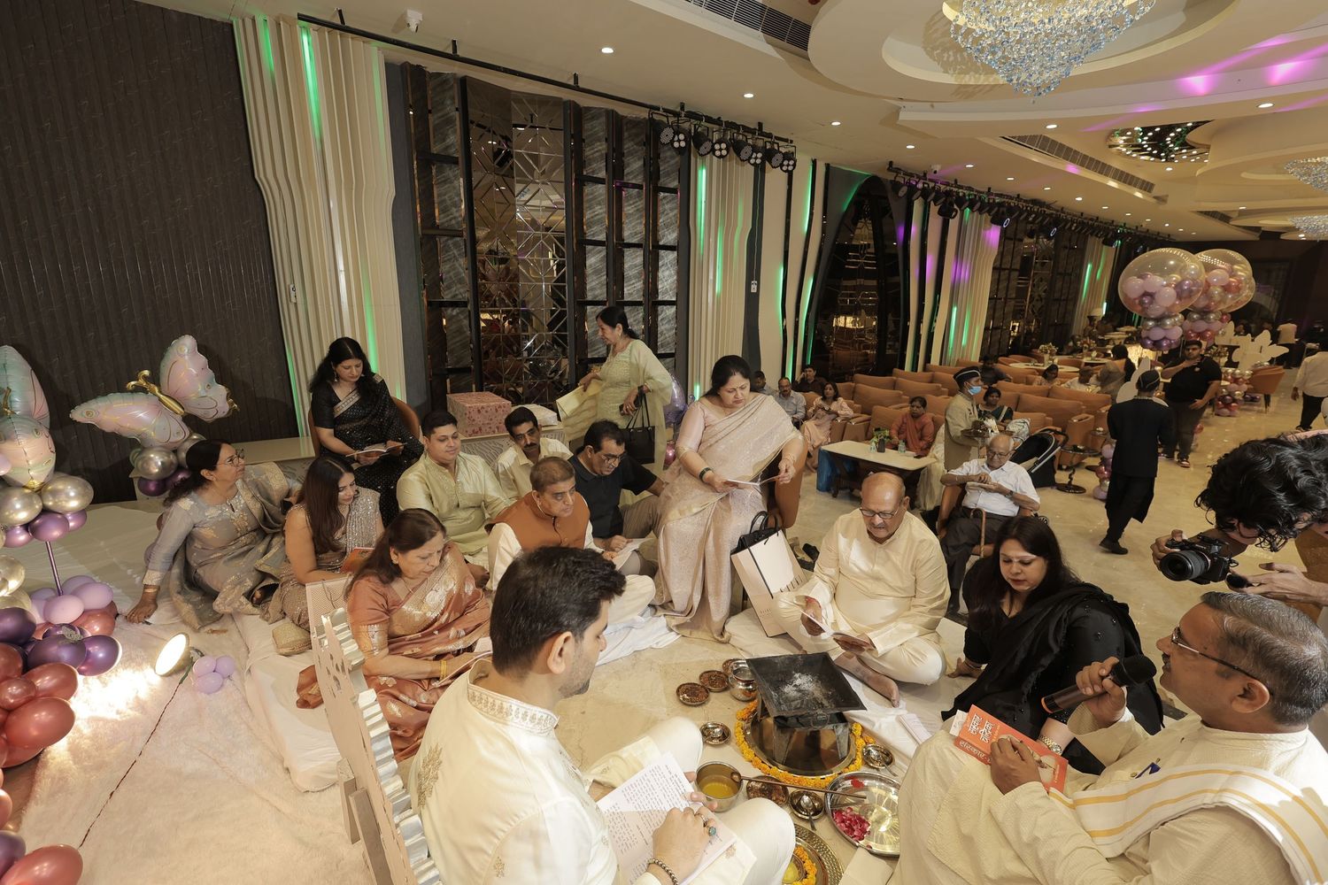 Family and guests gather on the floor of an elegant banquet hall for a traditional Indian ceremony with ritual items and balloon decor.