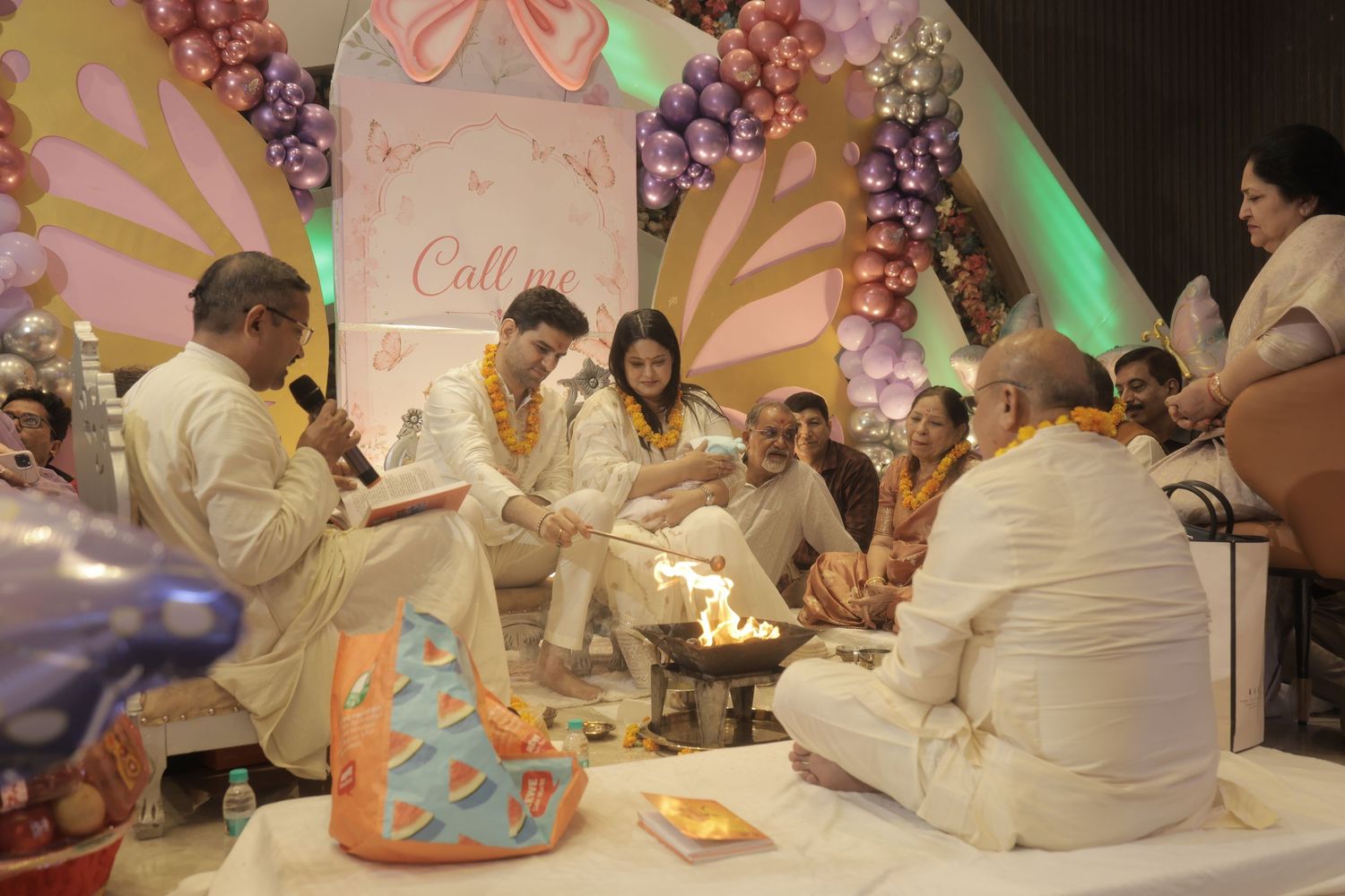Family members in traditional white attire sit around a sacred fire during a Hindu ceremony in a hall decorated with colorful balloons.