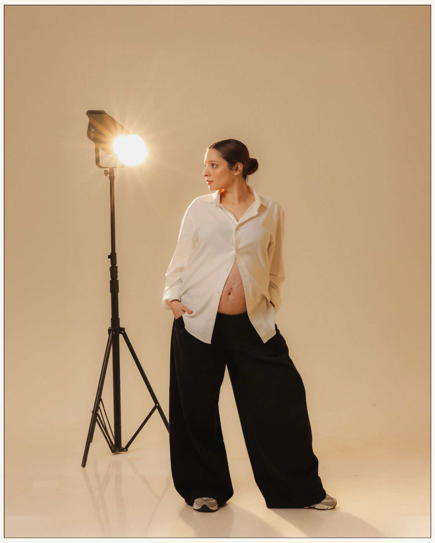 Pregnant woman in white shirt and black wide-leg pants poses beside a studio light on beige background.
