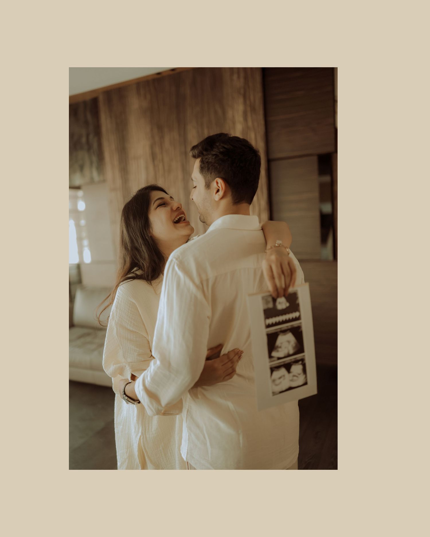 Couple in white embraces while looking at ultrasound photos in a warm, intimate indoor setting.