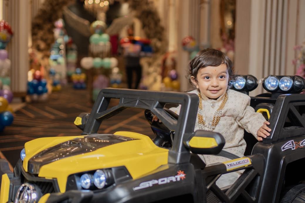 Young child in traditional attire smiling while riding a yellow toy SUV at a festive decorated event hall.
