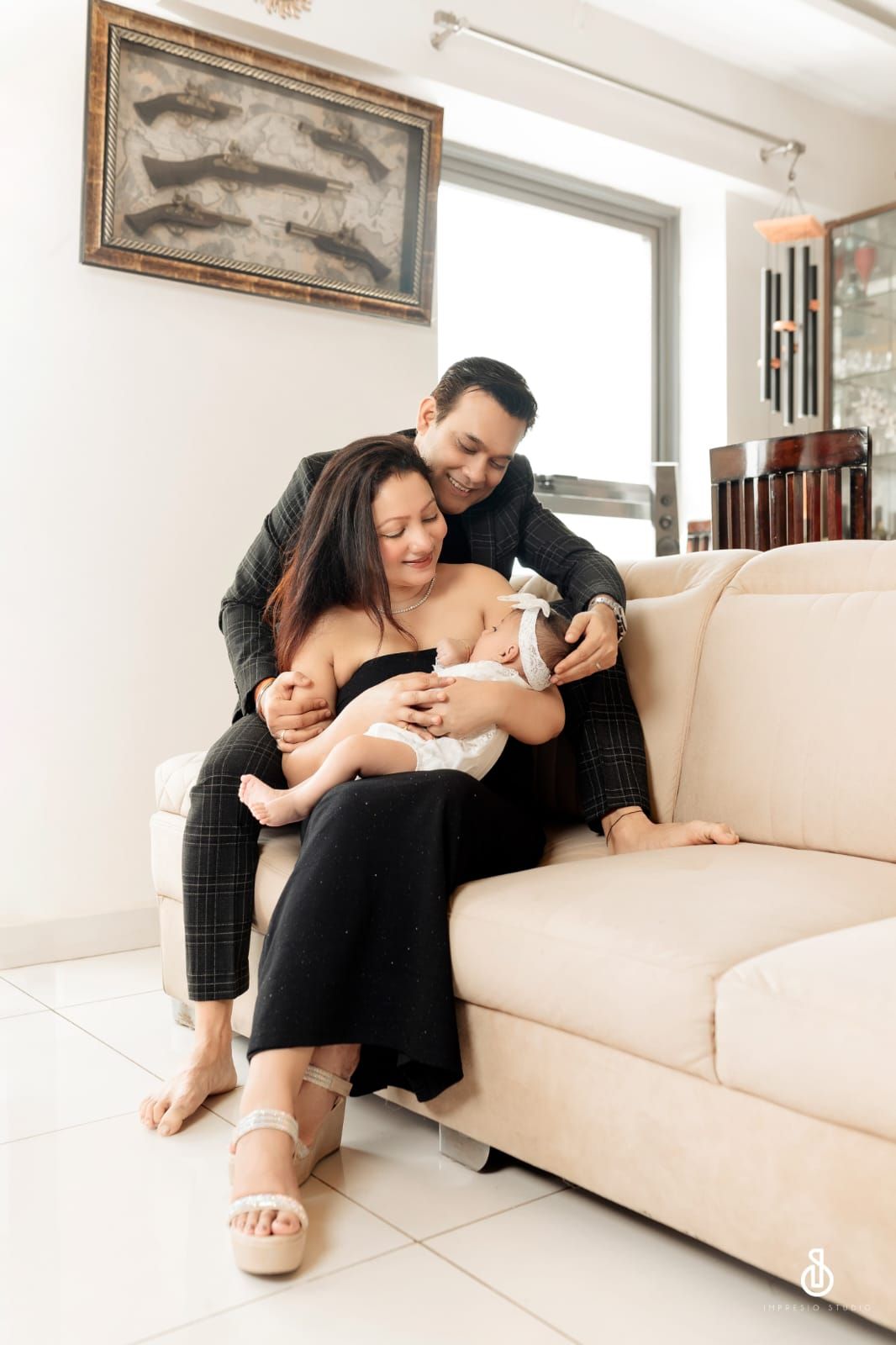 A couple sits on a beige sofa, lovingly cradling their newborn baby in a bright, modern living room.